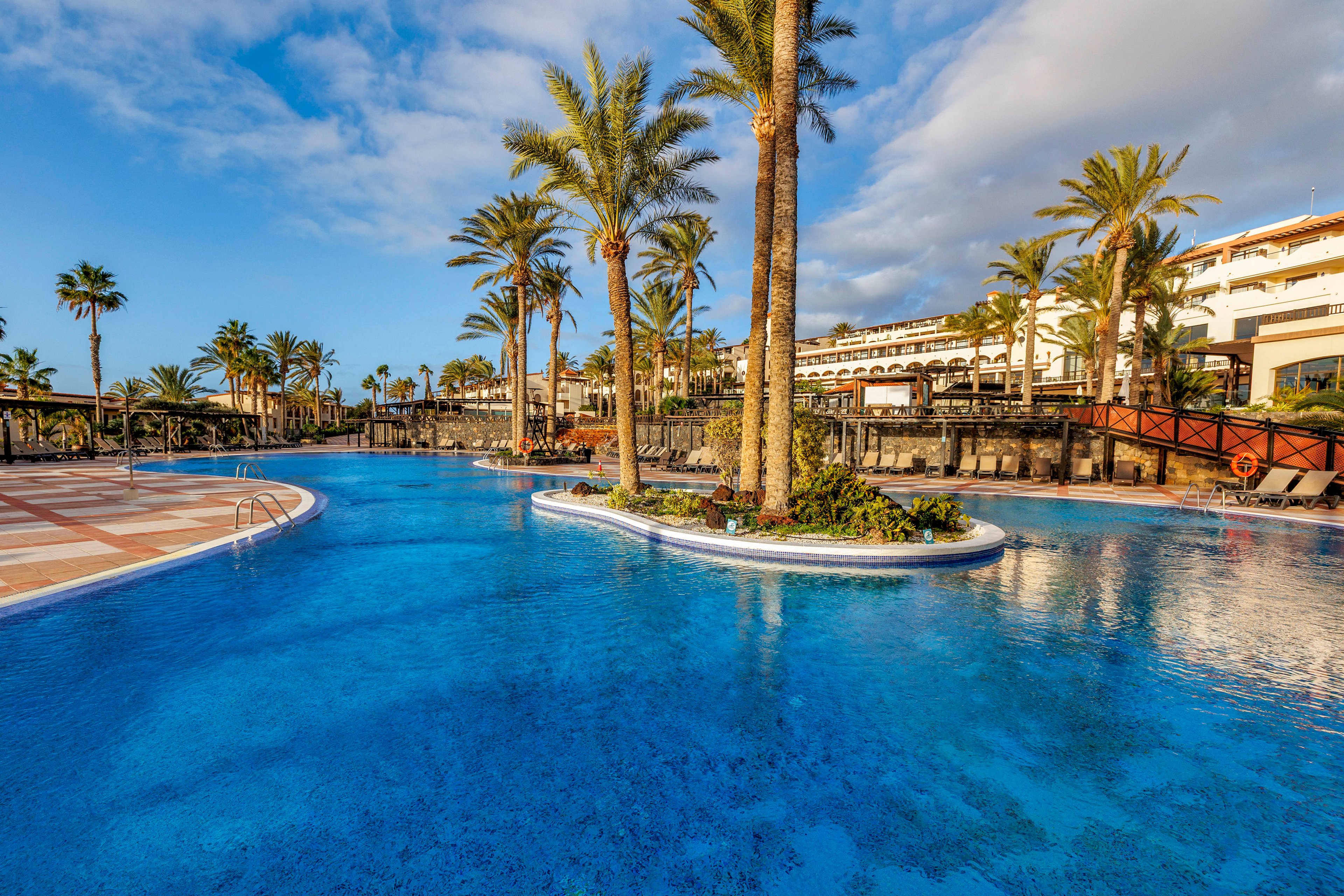 A large, curved swimming pool stretches across the foreground of a sunny resort scene. Tall palm trees line the pool, casting reflections on the clear blue water. A wooden bridge and landscaped pathways connect different areas of the resort buildings in the background. The bright sky with scattered clouds enhances the relaxed, vacation atmosphere.