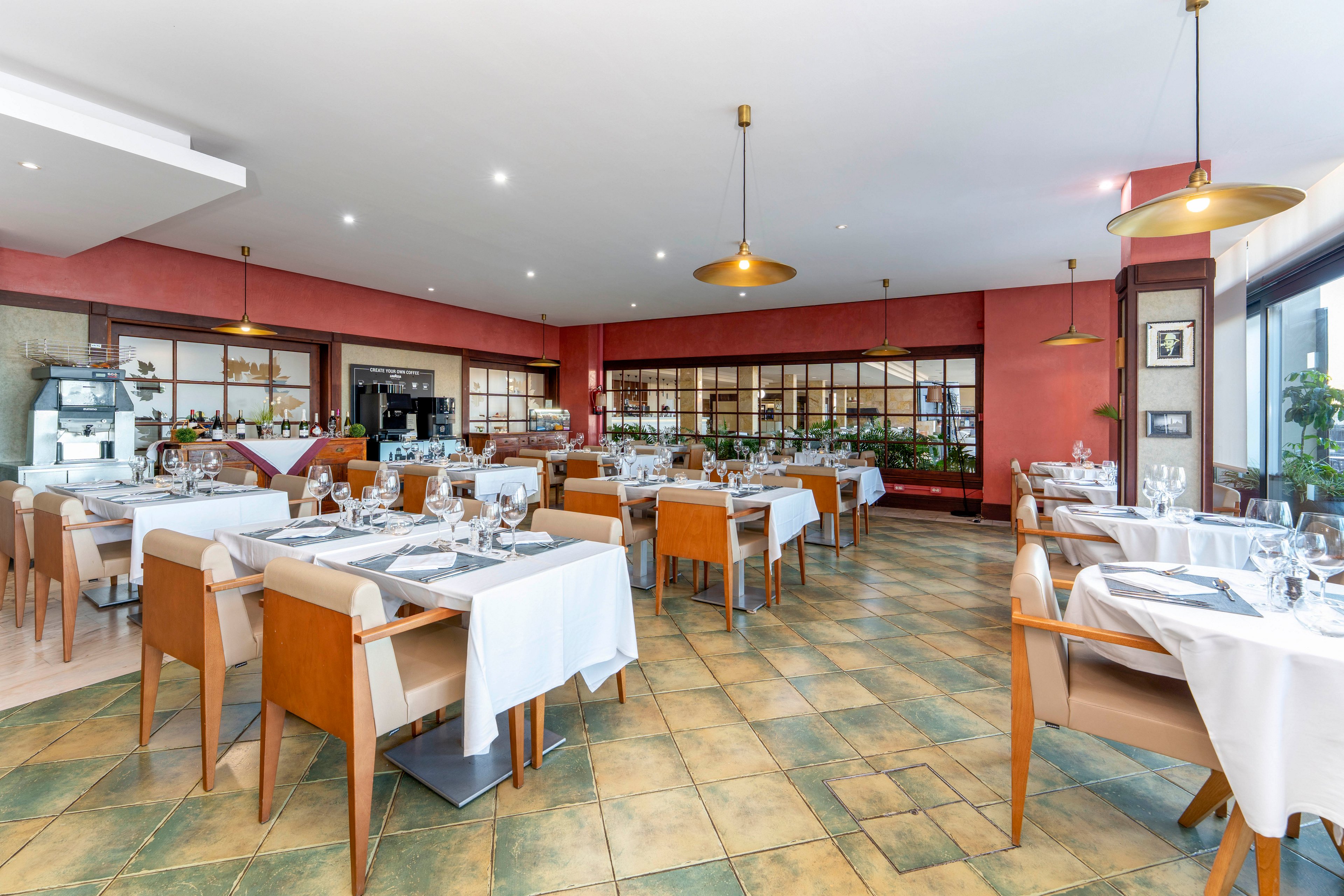 A contemporary restaurant dining room is set with neatly arranged tables and chairs. White tablecloths, glassware, and cutlery are prepared for guests, creating a formal yet inviting atmosphere. Warm lighting fixtures hang from the ceiling, illuminating the tiled floor and red accent walls. Large windows and glass doors allow natural light to brighten the spacious interior.
