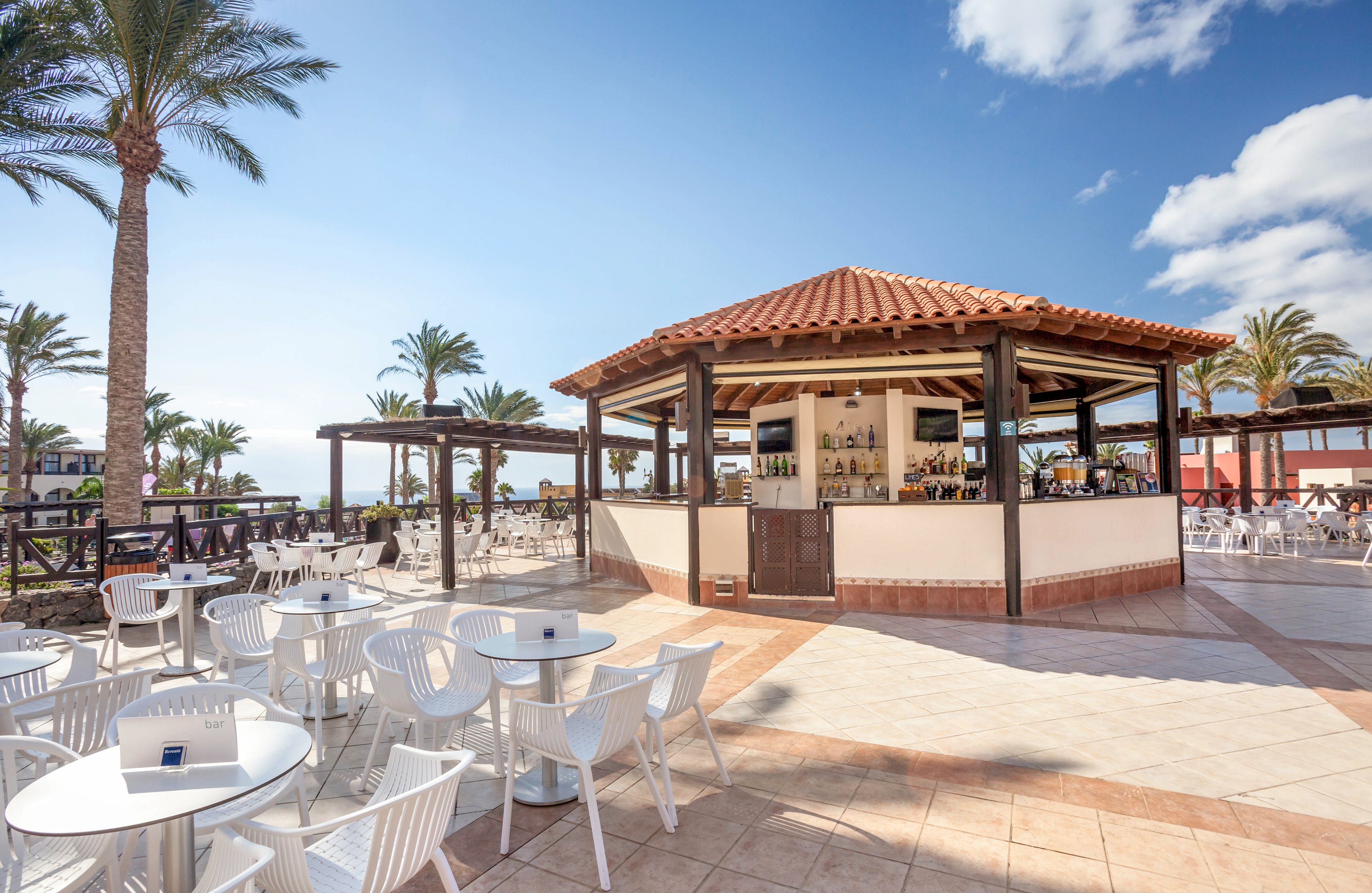 An open-air bar with a terracotta-tiled roof stands at the center of a spacious resort terrace. White tables and chairs are arranged around the bar, creating an inviting outdoor seating area. Palm trees, blue sky, and scattered clouds frame the sunny coastal setting. The scene feels relaxed and leisurely, ideal for vacation dining or drinks.