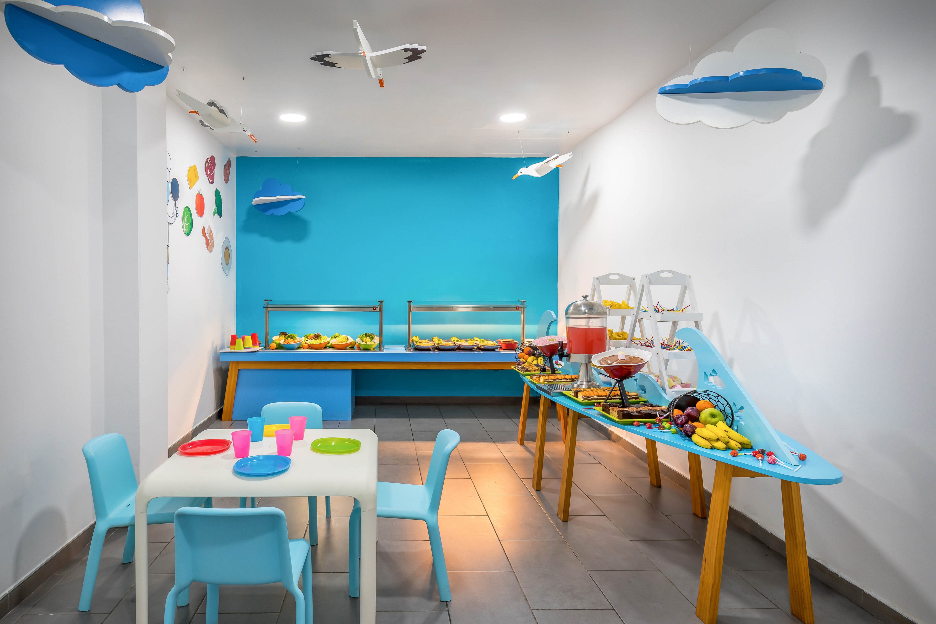 A bright kids buffet room features a small table with pastel-colored chairs and plastic cups in the foreground. Along the wall, a buffet counter and a long table display assorted fruits, pastries, and beverage dispensers. The walls are painted white with a bold blue accent wall, decorated with clouds, airplanes, and colorful circular decals. Overhead lighting and playful ceiling decorations create a cheerful, family-friendly atmosphere.