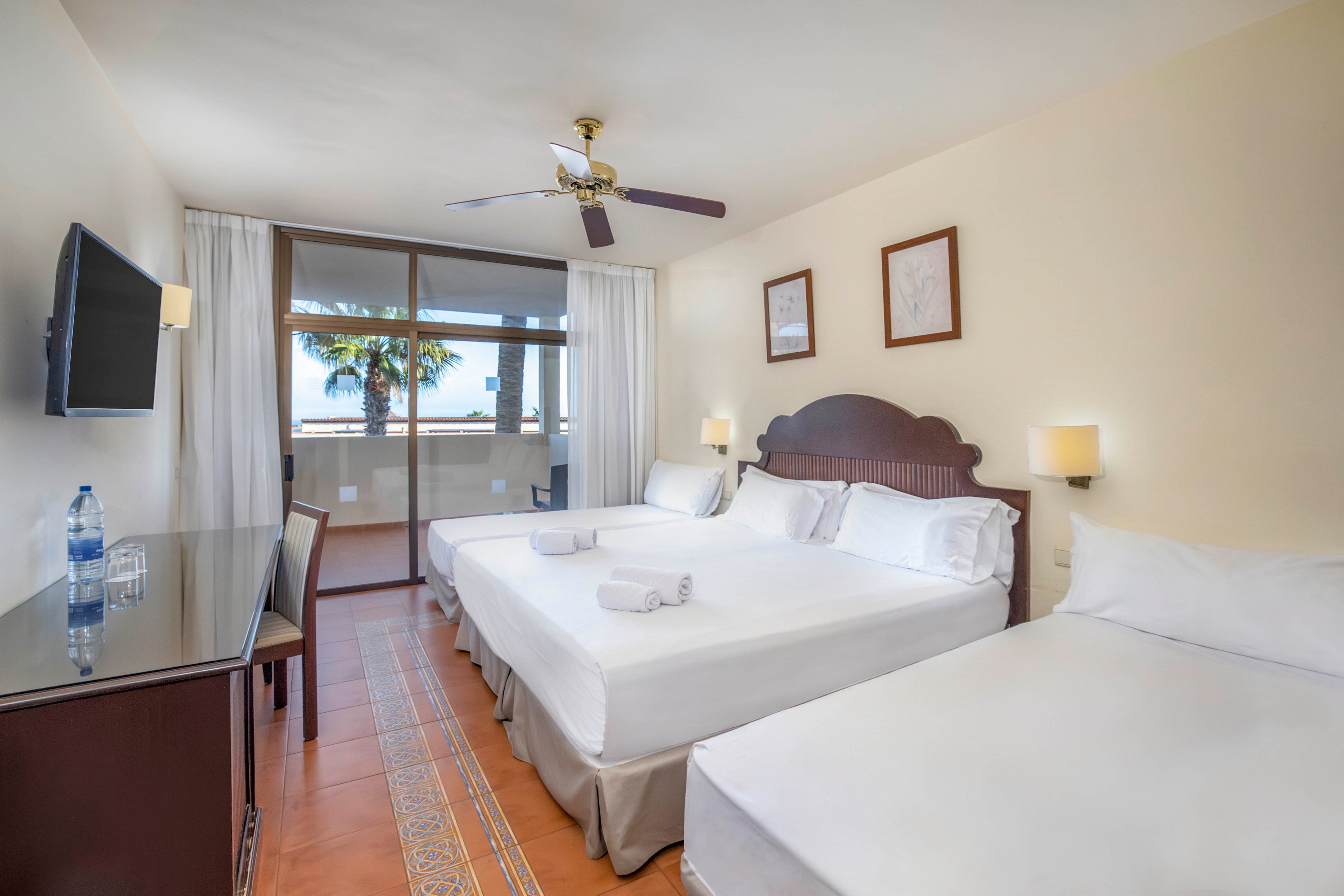 A bright hotel guest room features three neatly made beds with white linens and folded towels. The space includes a dark wooden desk with a flat-screen television mounted on the wall and a bottle of water placed on the surface. Large glass doors open to a balcony with a view of palm trees and the ocean in the background. Warm lighting, tiled floors, and neutral walls create a clean and inviting atmosphere.