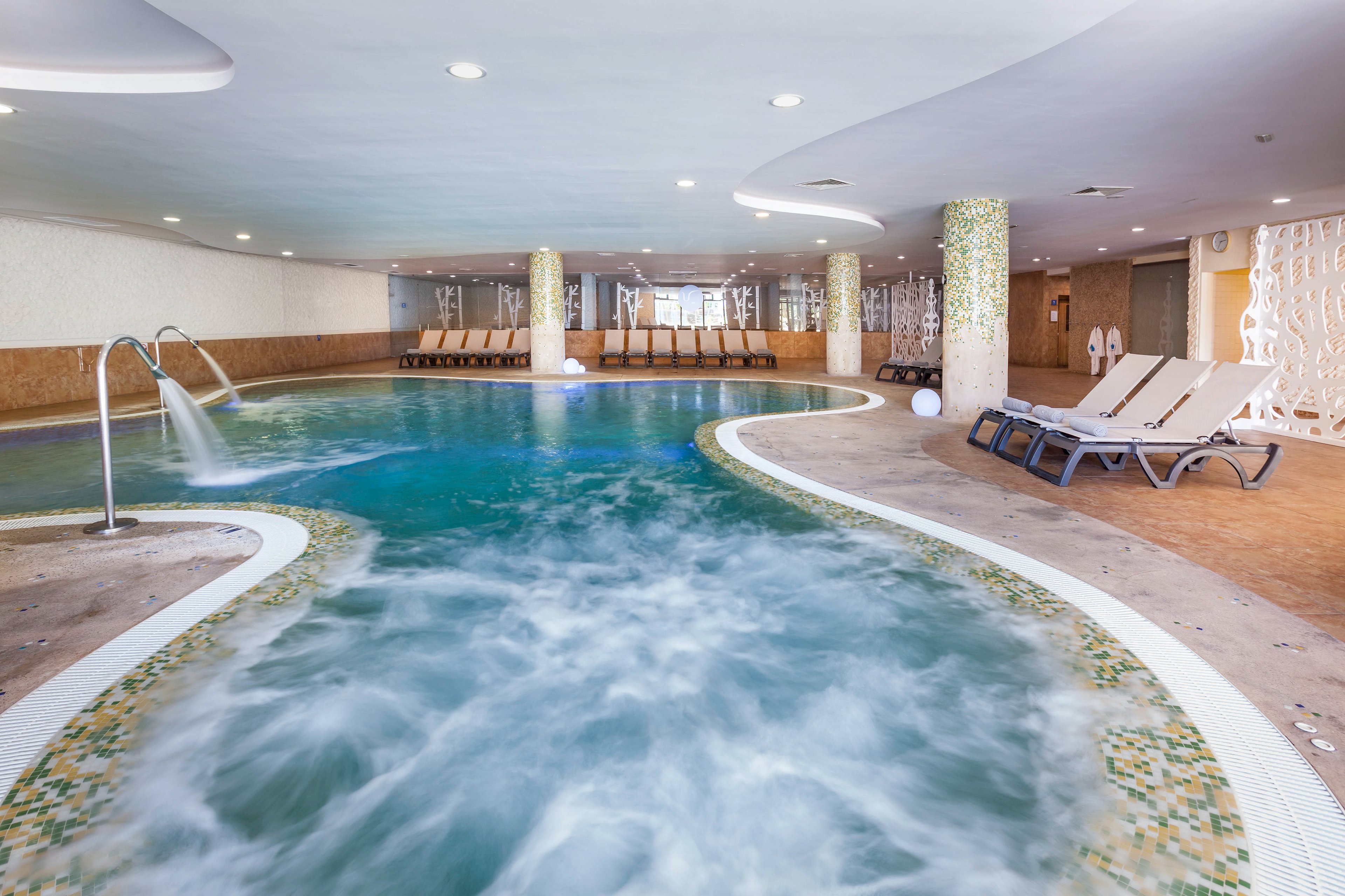 An indoor spa pool features swirling, bubbling water and a curved design. Lounge chairs line the far side of the pool, inviting guests to relax in the tranquil setting. Decorative columns and mosaic tile accents add a modern, upscale feel to the space. Soft lighting and neutral tones contribute to a calm, resort-like atmosphere.