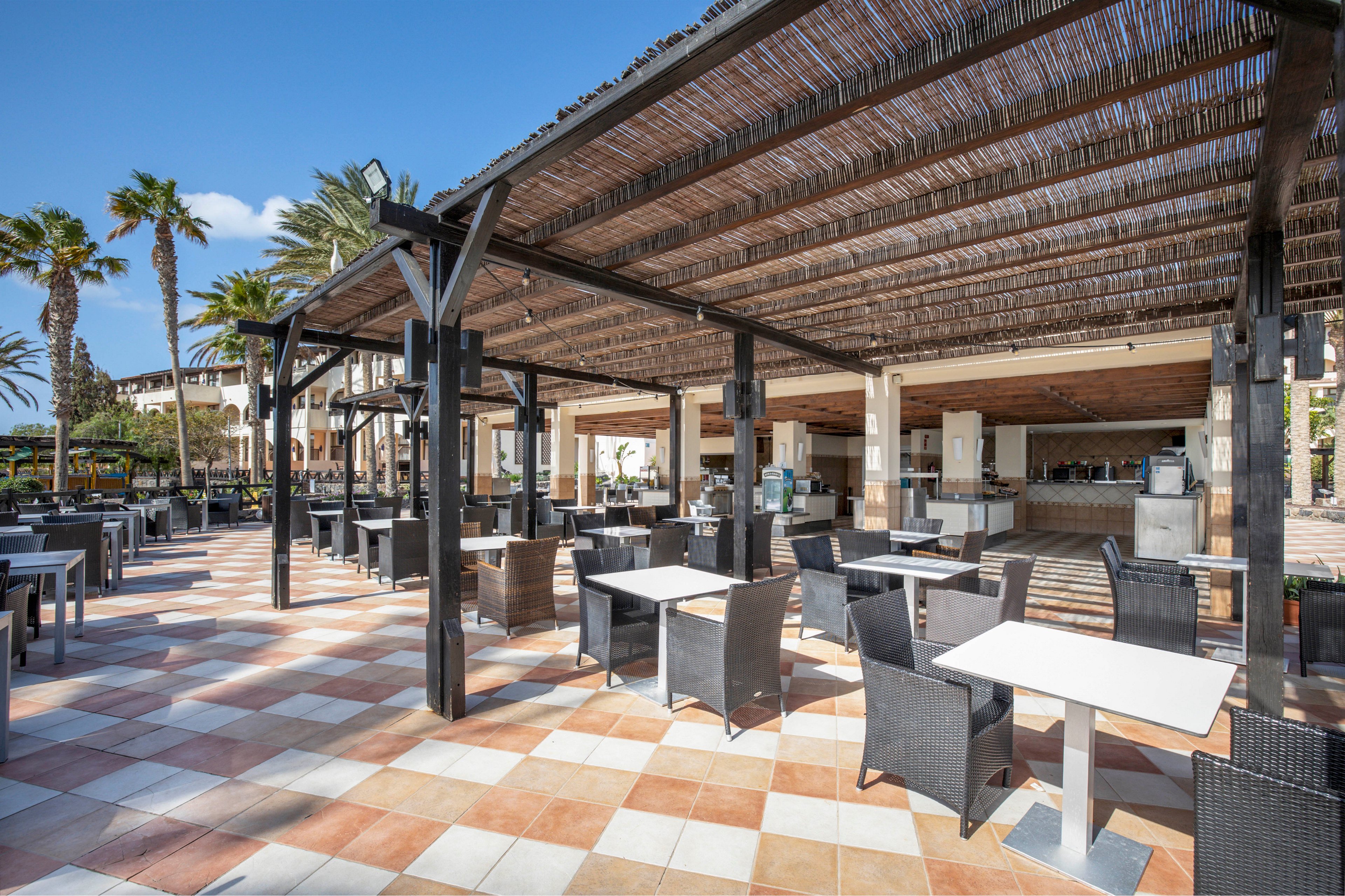 An open-air café terrace features rows of empty tables and dark wicker chairs under a wooden pergola. The space is part of a resort complex, with palm trees and additional buildings visible in the background. Sunlight filters through the slatted roof, casting shadows on the patterned tile floor. The scene feels calm and inviting, suggesting a relaxed vacation dining atmosphere.
