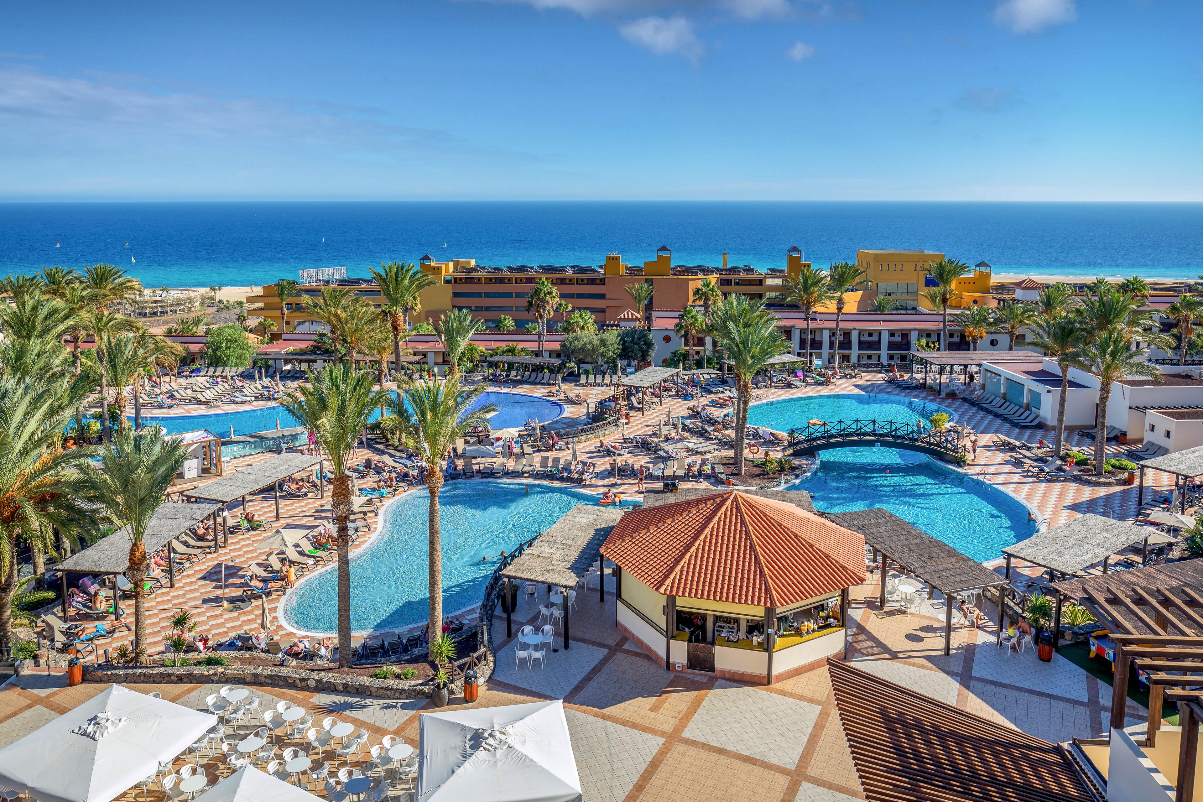 A large tropical resort features several bright blue swimming pools surrounded by palm trees and sun loungers. The complex overlooks a vivid turquoise ocean under a clear blue sky. Guests relax around the pools and walk along tiled pathways between buildings and shaded terraces. The scene conveys a sunny, coastal vacation atmosphere with abundant outdoor leisure spaces.