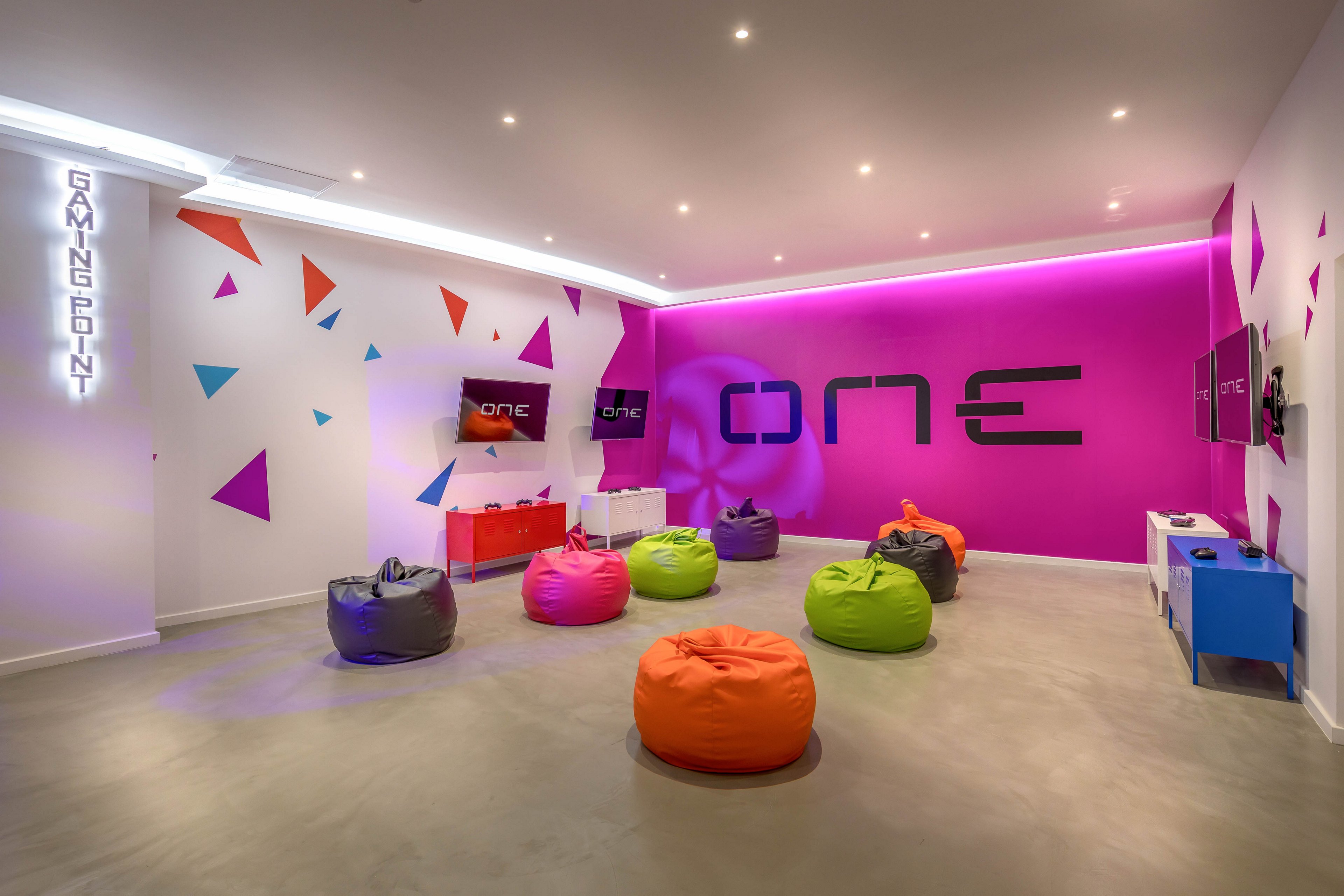 A modern gaming lounge features a row of colorful bean bag chairs arranged in the center of the room. The space has bright white walls accented with multicolored geometric shapes and a vivid magenta feature wall. Large wall graphics display the word "one" alongside mounted screens and gaming consoles. Recessed ceiling lights create an evenly lit, contemporary atmosphere suited for social gaming or relaxation.