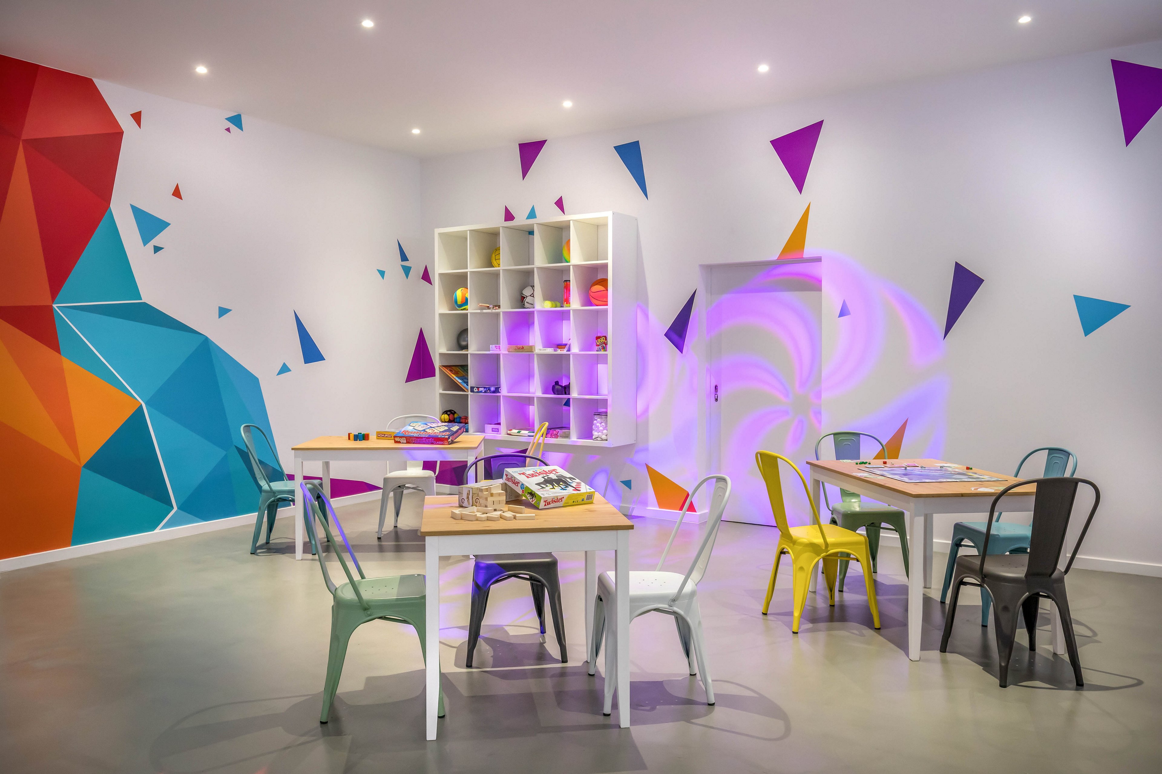 A bright, modern classroom features several tables with colorful metal chairs arranged around them. The walls are decorated with bold geometric murals and abstract shapes in vibrant hues. A cube-shelving unit at the back holds various supplies and toys, suggesting a space for art, crafts, or creative learning. Recessed ceiling lights and soft accent lighting create a playful yet organized atmosphere suitable for children or group activities.
