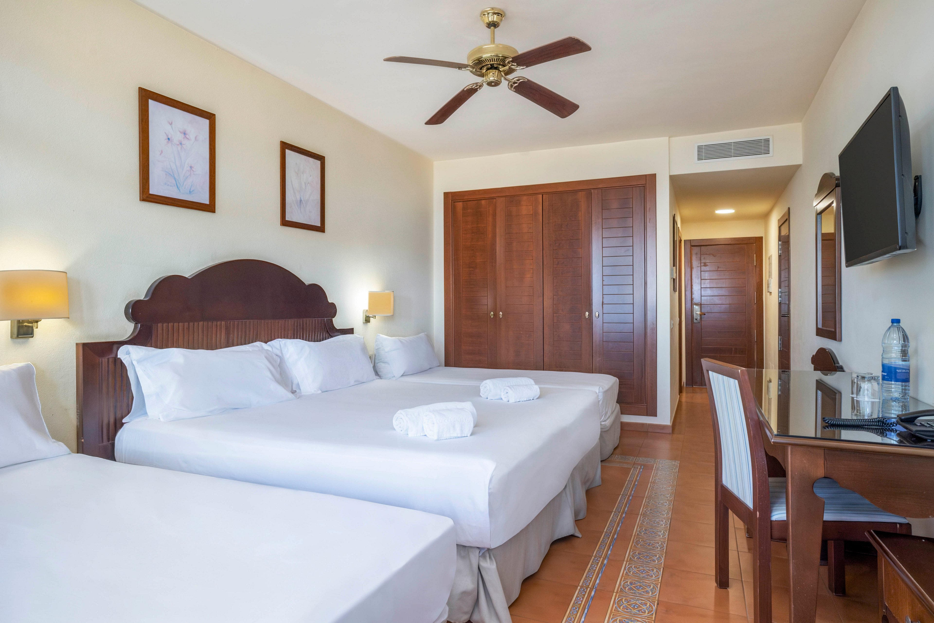 A bright hotel guest room features three neatly made beds with white linens and folded towels placed on top. Warm wooden furniture, including a large wardrobe and desk, contrasts with the light-colored walls and tiled floor. A ceiling fan and wall-mounted television add comfort and convenience for guests. Soft lighting and framed artwork create a clean, welcoming atmosphere suitable for families or groups.
