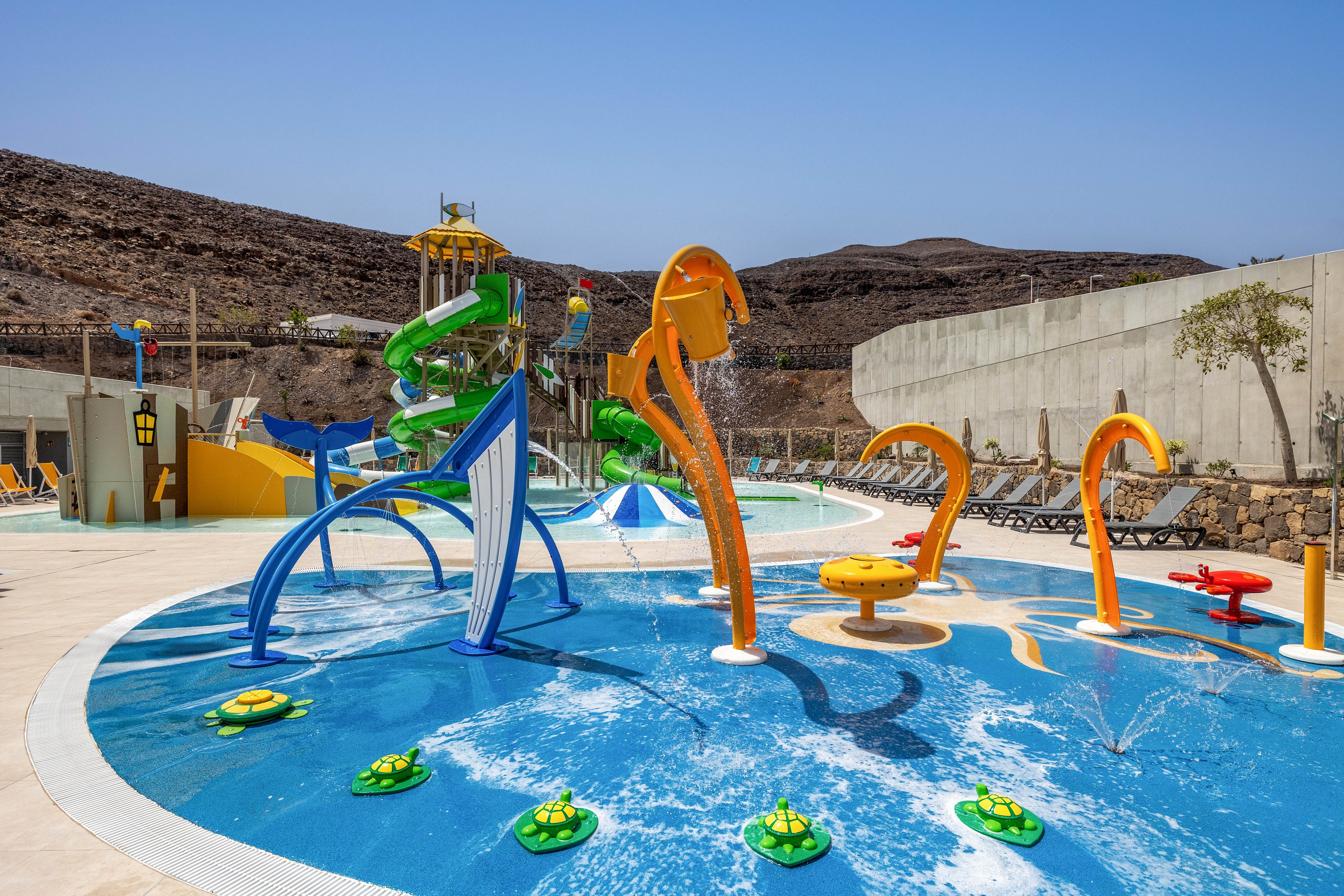 A vibrant outdoor splash park features colorful water play structures and fountains. Curved orange and blue sprayers arch over a shallow pool with interactive elements shaped like flowers and sea creatures. In the background, tall twisting water slides rise against a rocky hillside under a clear blue sky. Lounge chairs line the perimeter, suggesting a family-friendly recreational area.