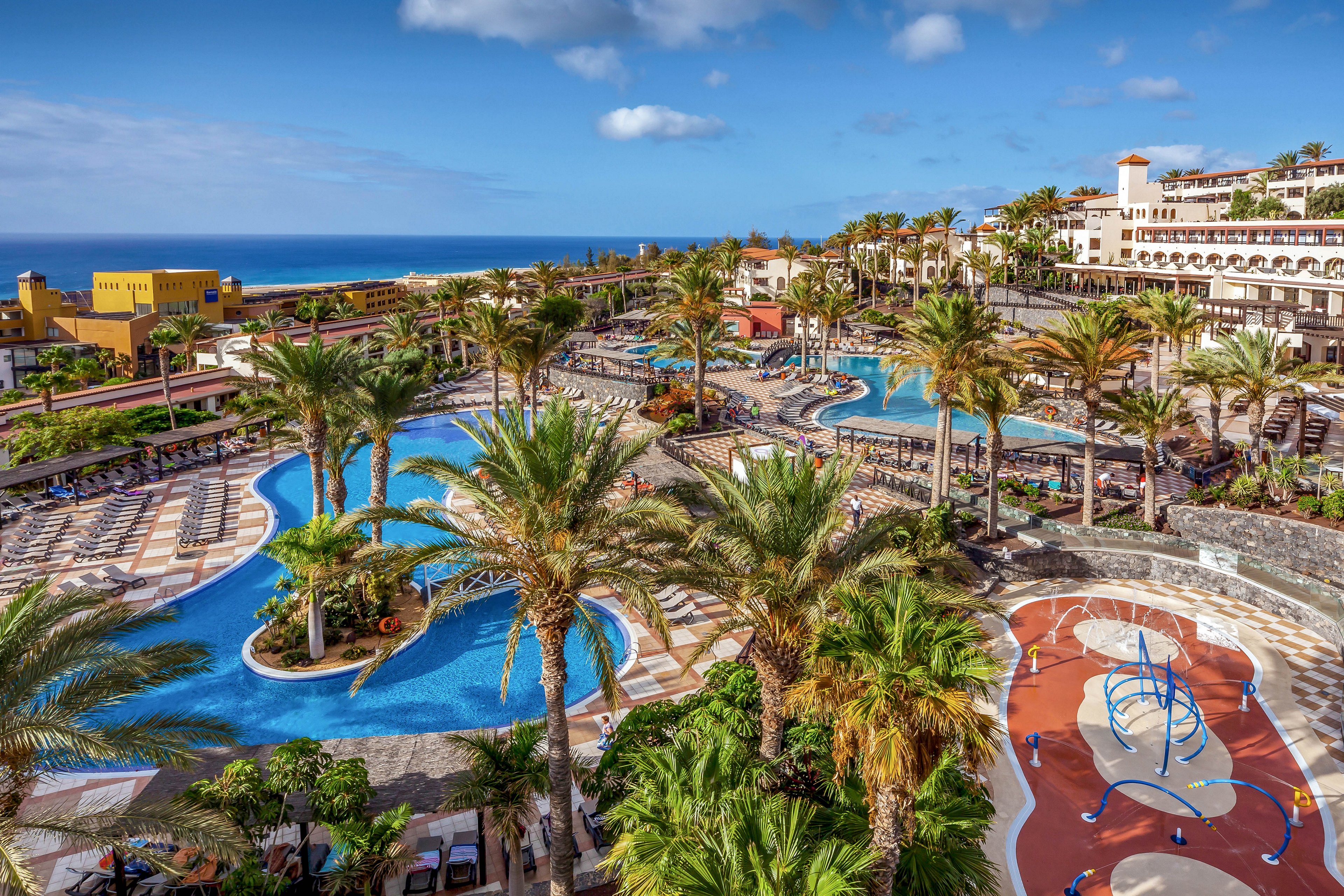 A large tropical resort features multiple curving swimming pools surrounded by palm trees and sun loungers. The complex includes a colorful playground area with a blue climbing structure and patterned ground. Multi-story hotel buildings line the background, overlooking the resort grounds. In the distance, the blue ocean and partly cloudy sky create a scenic coastal backdrop.