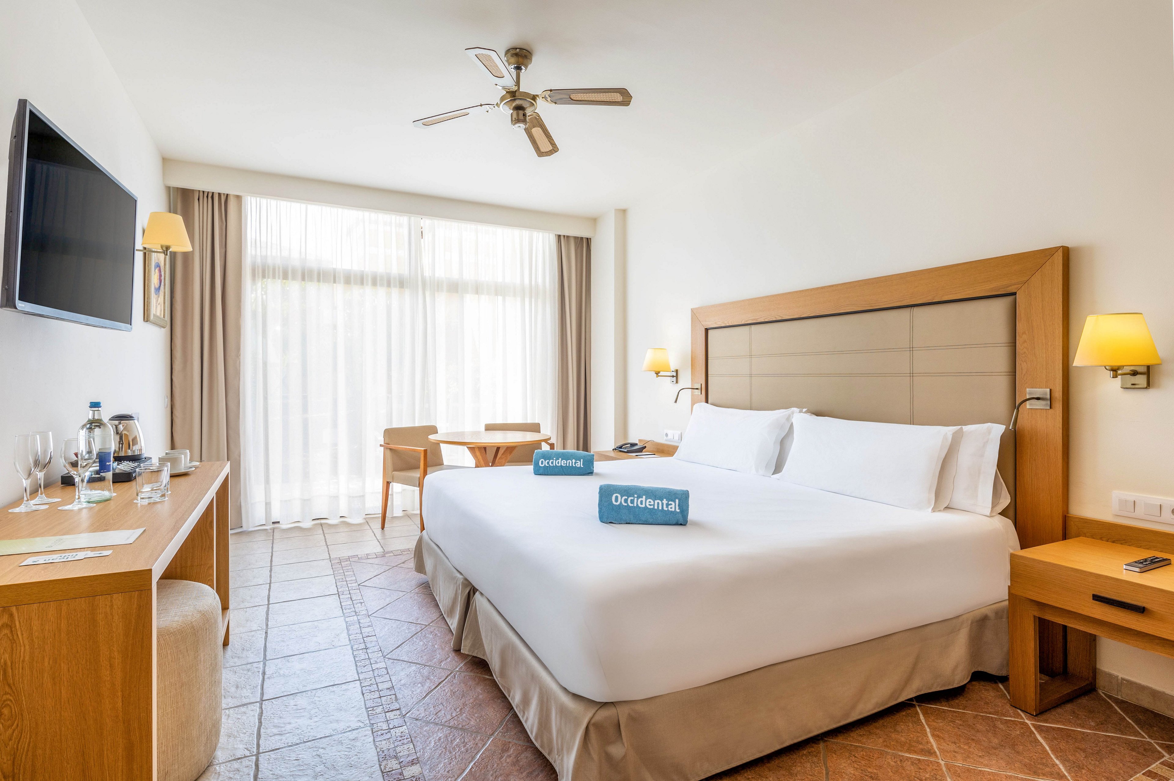 A spacious hotel room features a large neatly made bed with white linens and a padded headboard. Natural light floods in through floor-to-ceiling curtains covering a wide window or balcony door. A wooden desk with bottled water, glasses, and a flat-screen TV contrasts with the tiled floor and neutral walls. Warm bedside lamps and a ceiling fan add comfort to the clean, contemporary space.