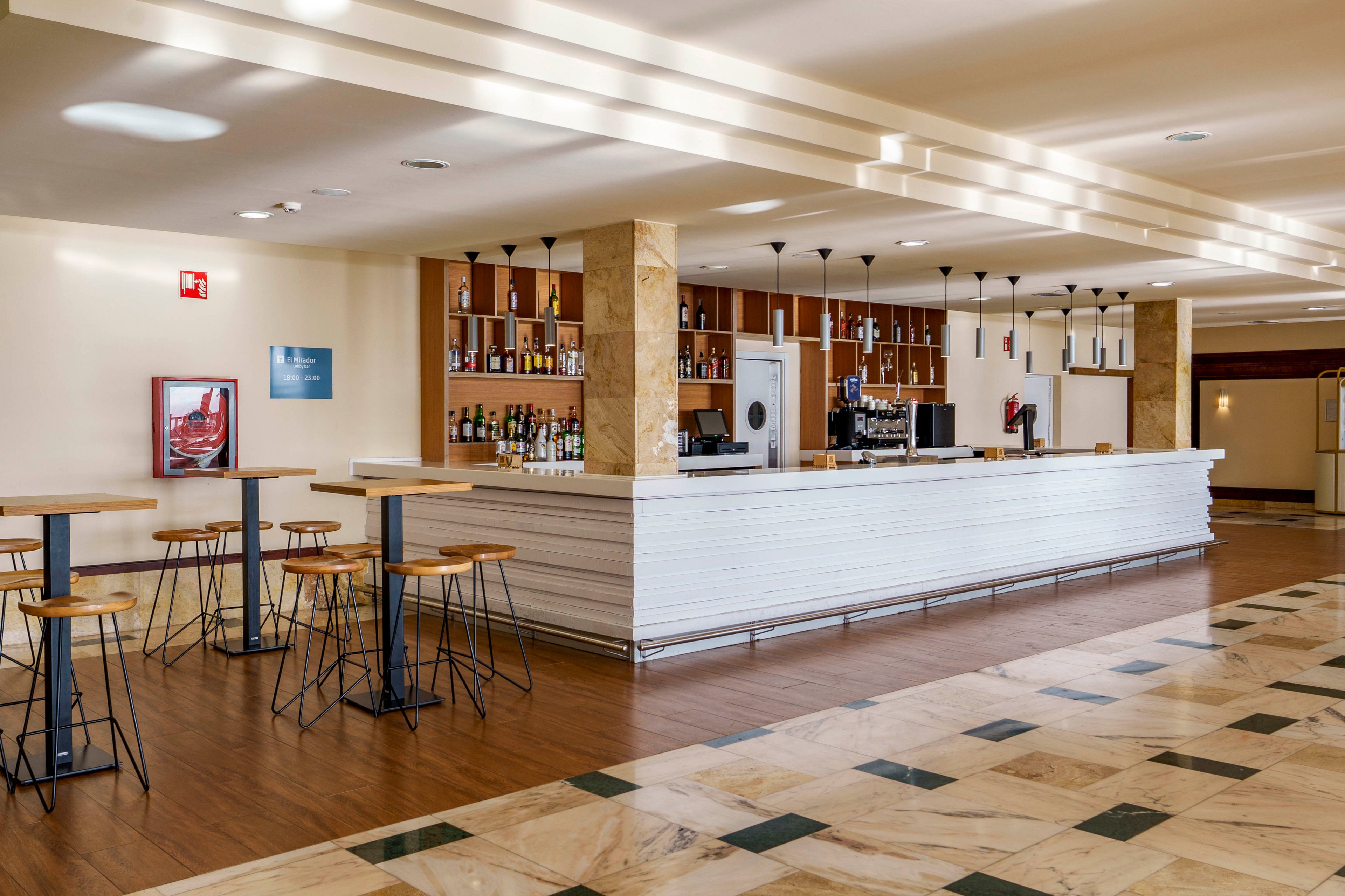 A contemporary hotel bar features a long white counter lined with bottles and glassware. Tall wooden bar stools and high tables are arranged neatly on a polished wooden floor. The space is empty of people, highlighting the clean lines and minimalist design. Warm lighting and neutral tones create an inviting, relaxed atmosphere.