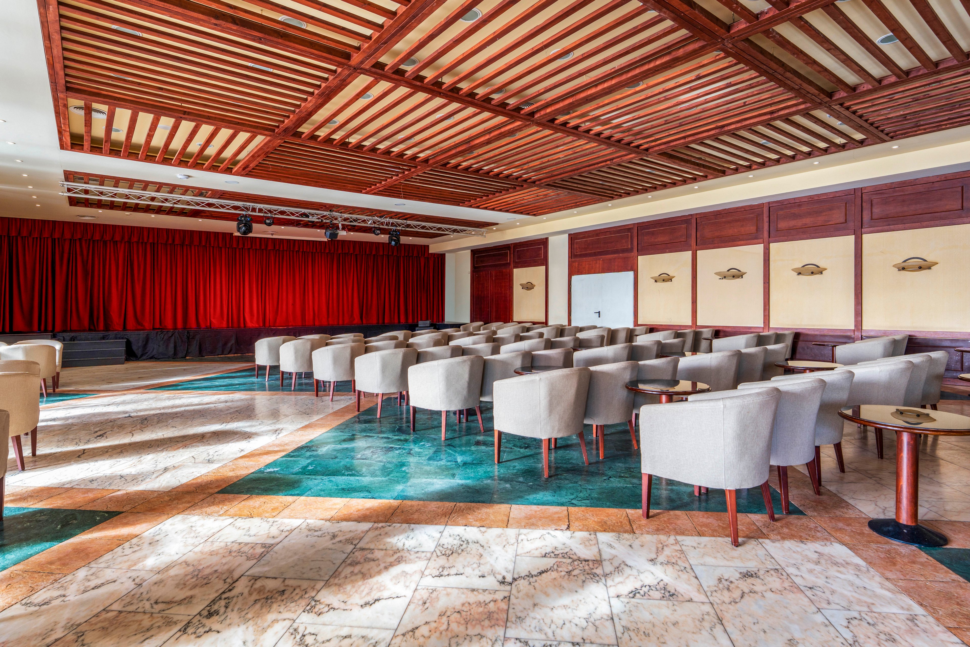 A spacious conference room is arranged with rows of light upholstered armchairs facing a stage. The stage features a long red curtain and overhead lighting equipment. The room has a patterned marble floor with green accents and wooden wall panels. A decorative wooden slatted ceiling adds warmth and texture to the interior, while small side tables accompany some of the chairs.