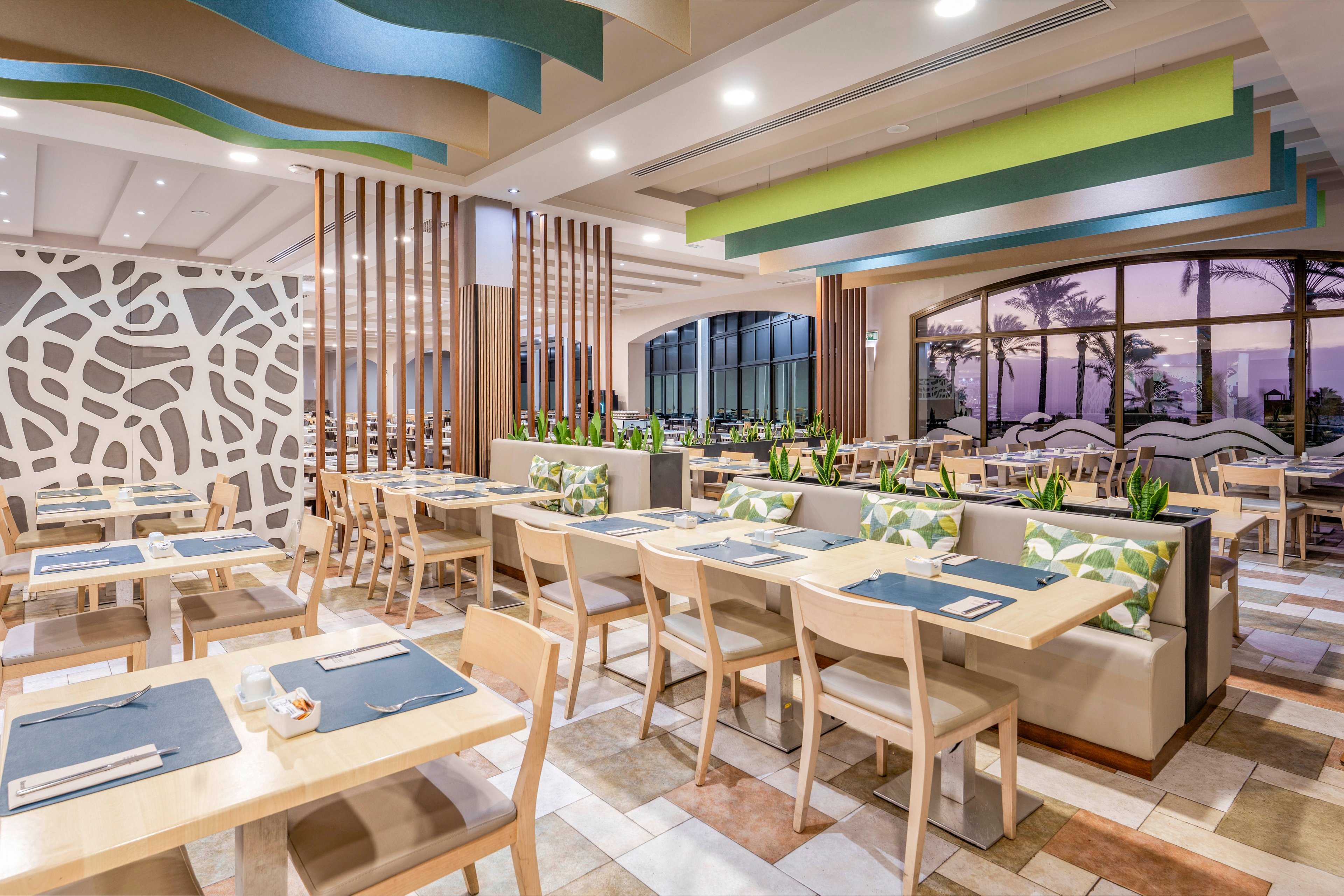 A contemporary restaurant dining area features light wooden tables and chairs arranged neatly in an open-plan layout. The space is decorated with geometric wall patterns, vertical wooden slats, and built-in banquette seating with patterned cushions. Large windows reveal palm trees and a colorful sunset sky outside, adding a coastal ambiance. Soft ceiling lighting and pastel accents in blue and green create a bright, inviting atmosphere.