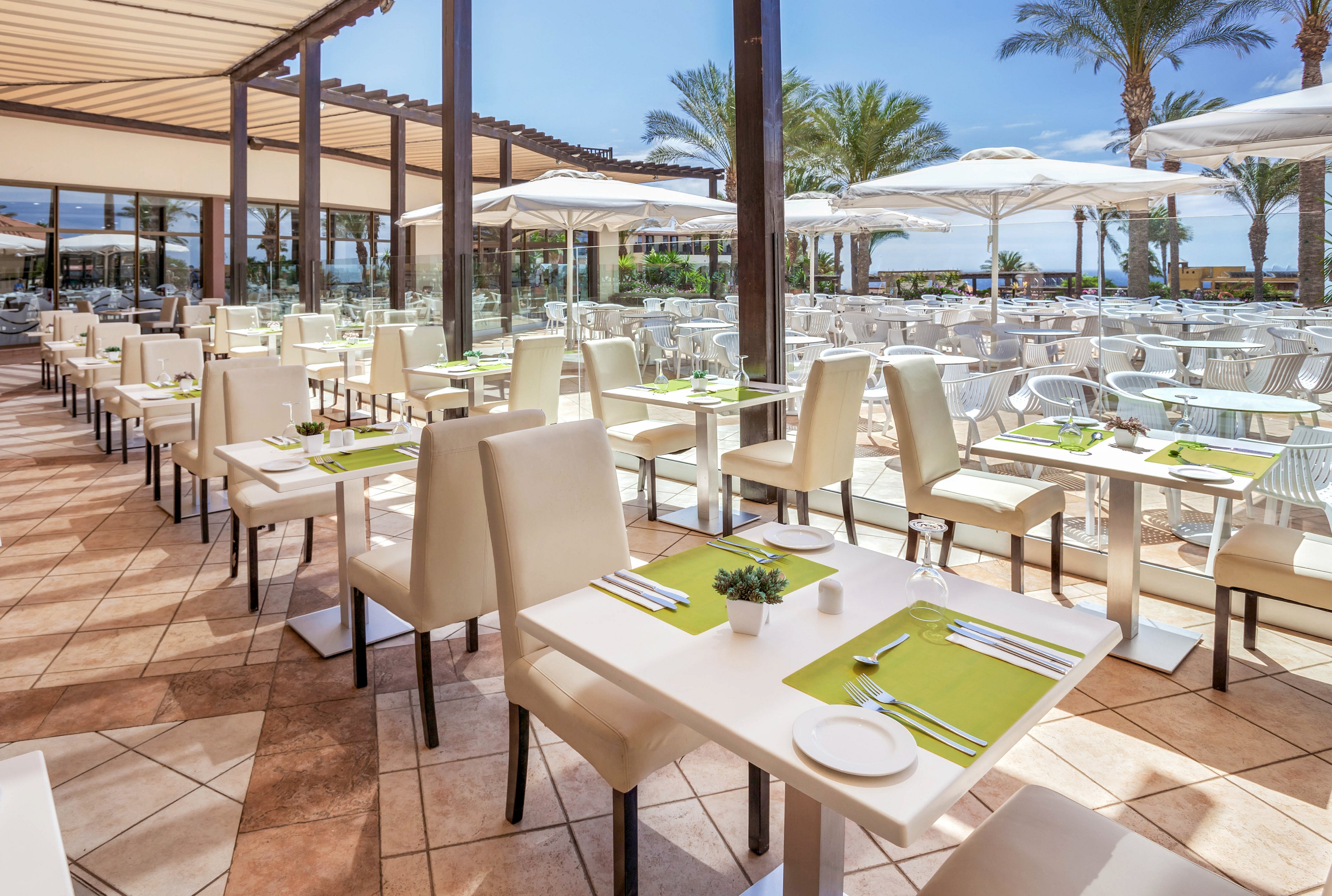 A contemporary restaurant terrace features neatly arranged white tables and cream-colored chairs. Each table is set with green placemats, cutlery, and small potted plants, creating a fresh and inviting dining atmosphere. Large glass panels separate the covered seating area from the open-air patio with white umbrellas and metal chairs. Palm trees and a distant view of the sea enhance the relaxed, resort-like setting under a bright blue sky.