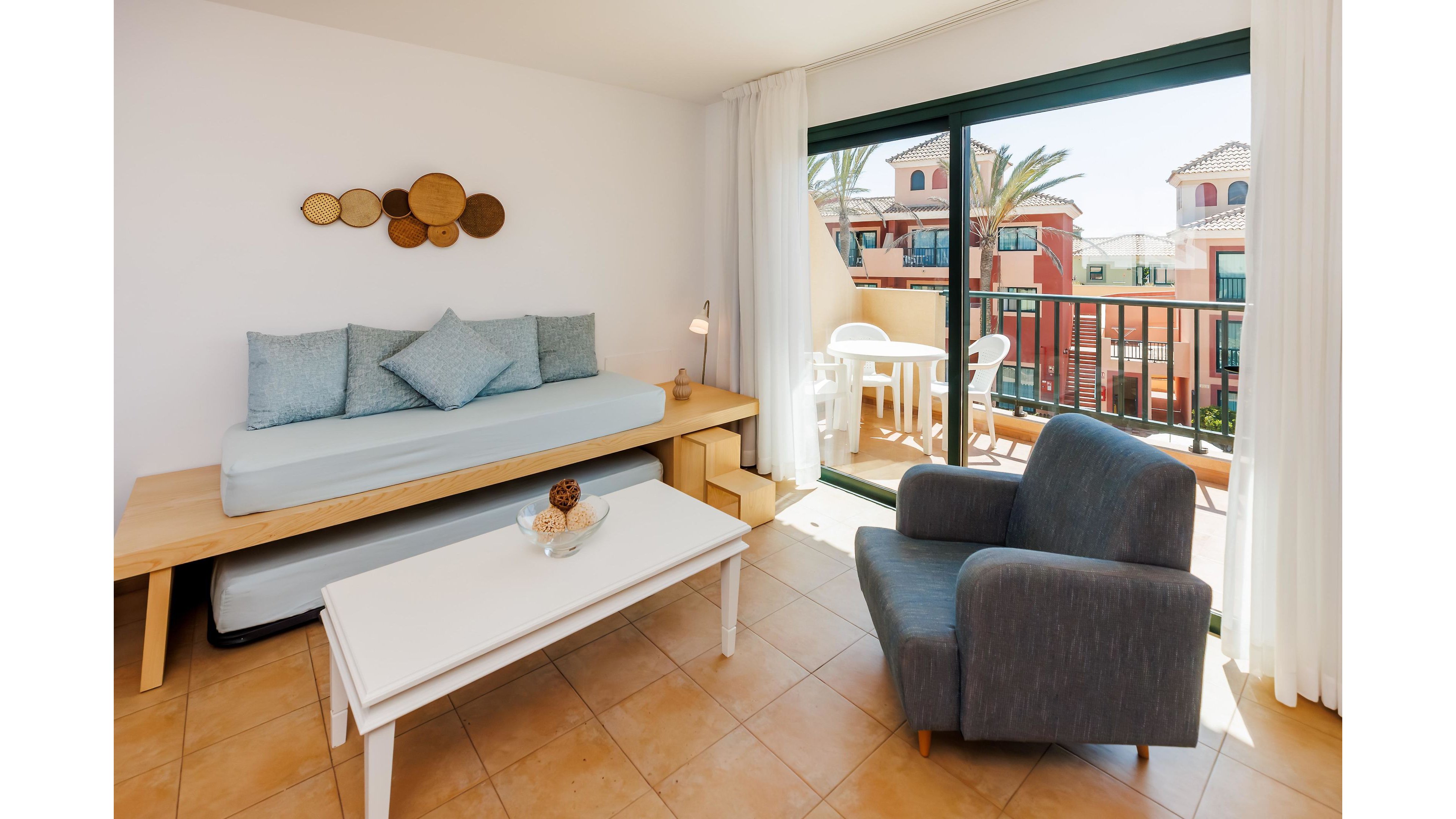 Sunlit living room in a vacation apartment features a minimalist daybed with light blue cushions and a modern gray armchair. A white coffee table with a small decorative bowl sits on a tiled floor, creating a clean and airy interior. Large sliding glass doors open to a balcony with outdoor chairs and a table, overlooking colorful Mediterranean-style buildings and palm trees. Neutral tones and simple decor give the space a relaxed, contemporary feel.