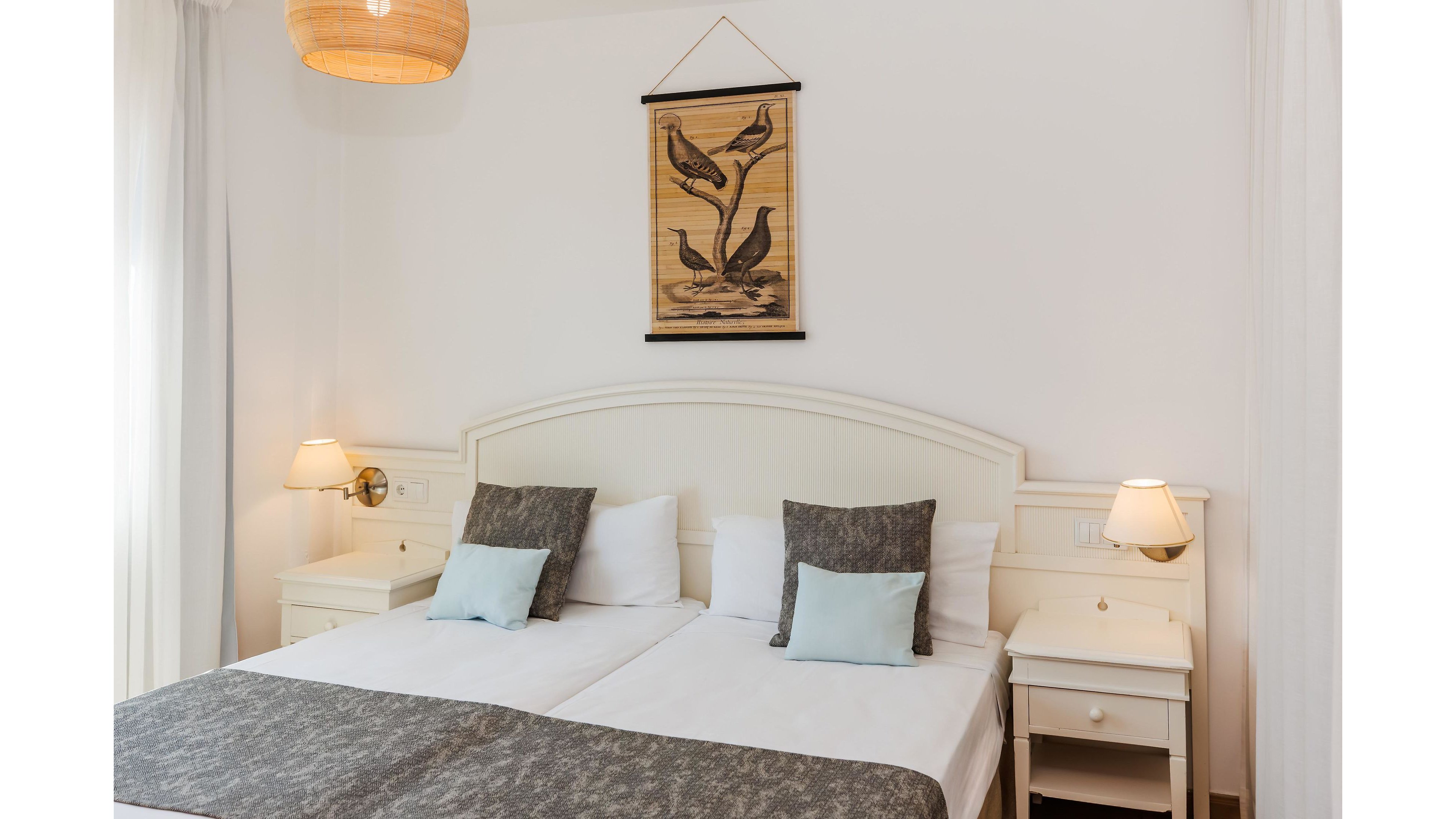 A bright, cozy bedroom features two neatly made twin beds with white linens and gray throws. Soft pastel cushions add a touch of color against the light headboard and matching bedside tables. Warm lighting from wall sconces and table lamps creates a welcoming atmosphere. A woven hanging artwork and natural pendant lamp enhance the simple, modern decor.
