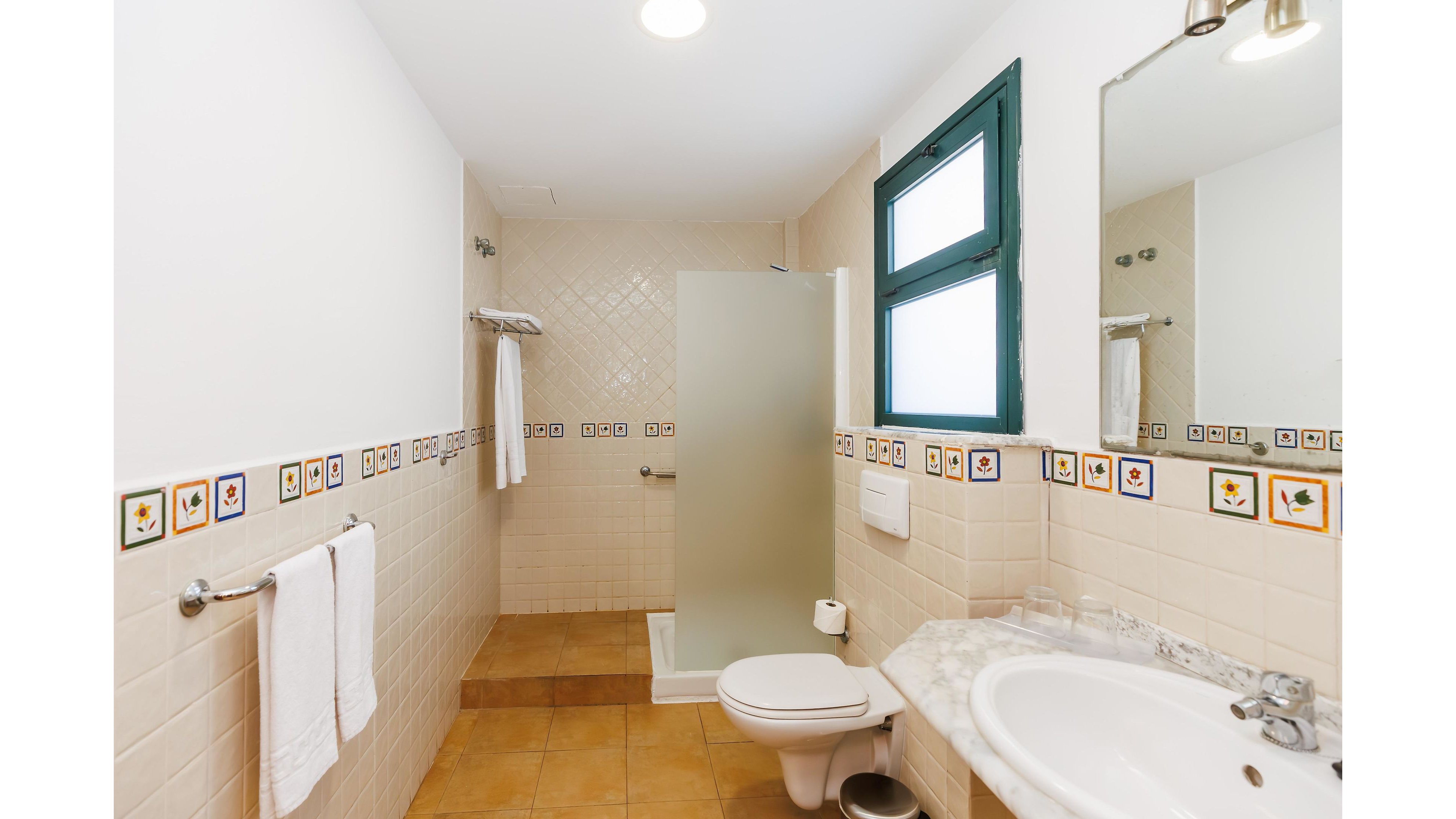 Clean, bright bathroom interior featuring a walk-in shower with frosted glass panel and a white toilet. A white sink with marble-style countertop sits to the right beneath a large wall mirror and overhead lights. Decorative border tiles with colorful motifs run along the walls above beige wall tiles and terracotta floor tiles. Towels hang neatly on chrome towel bars, and a green-framed window adds natural light to the space.
