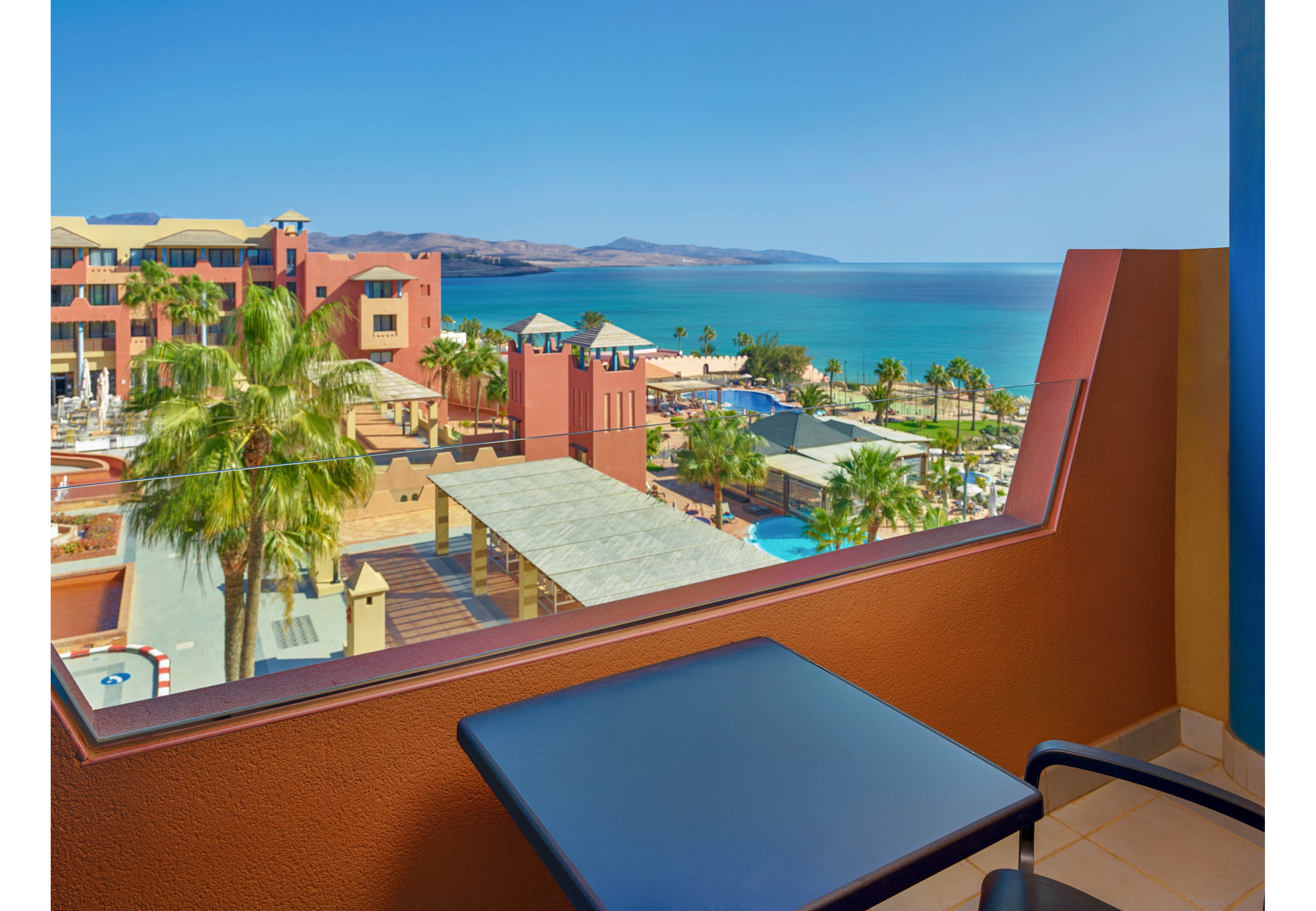 A resort balcony overlooks a coastal hotel complex with palm trees and bright terracotta buildings. In the foreground, a small table holds two glasses of orange juice and a plate of sliced fruit. Beyond the balcony, guests can see a swimming pool area, walkways, and the blue ocean stretching to distant hills. The scene is bathed in clear daylight, creating a relaxed vacation atmosphere.