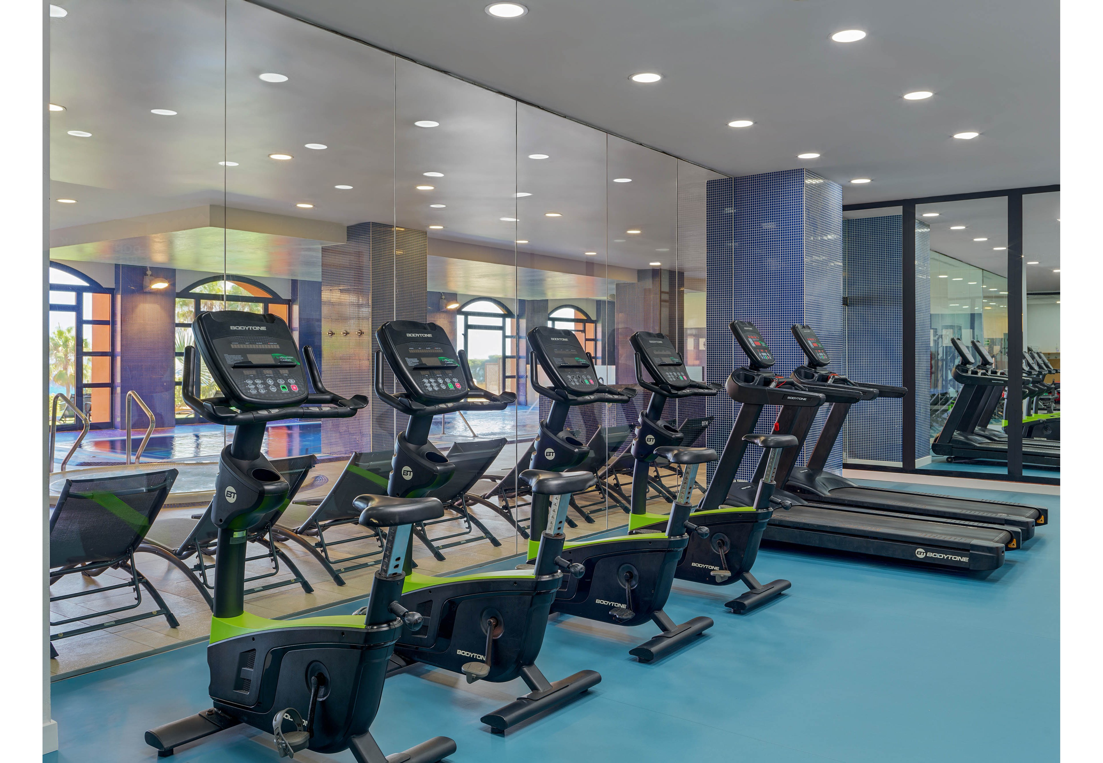 A contemporary indoor gym features a row of stationary bikes and treadmills neatly aligned on a blue floor. Large mirrors along the wall reflect the machines and the bright, spacious interior. In the background, lounge chairs and arched windows suggest an adjacent pool or relaxation area. Recessed ceiling lights and clean lines create a sleek, professional fitness environment.