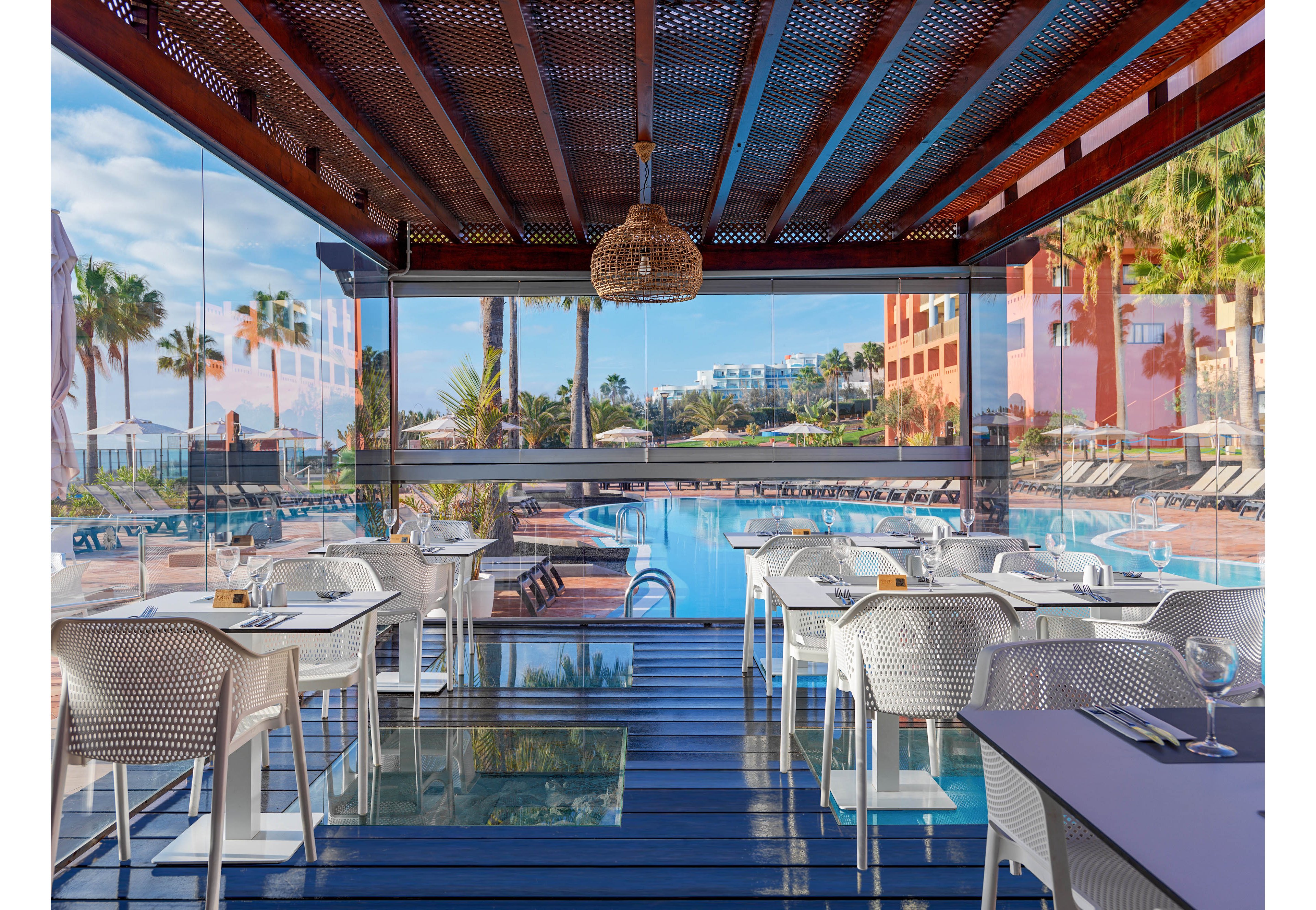 An open-air restaurant terrace overlooks a large resort swimming pool. White tables and chairs are neatly arranged on a deck with glass floor panels. Glass walls frame views of palm trees, sun loungers, and colorful hotel buildings under a bright sky. The scene feels calm and inviting, suggesting a relaxed vacation dining experience.