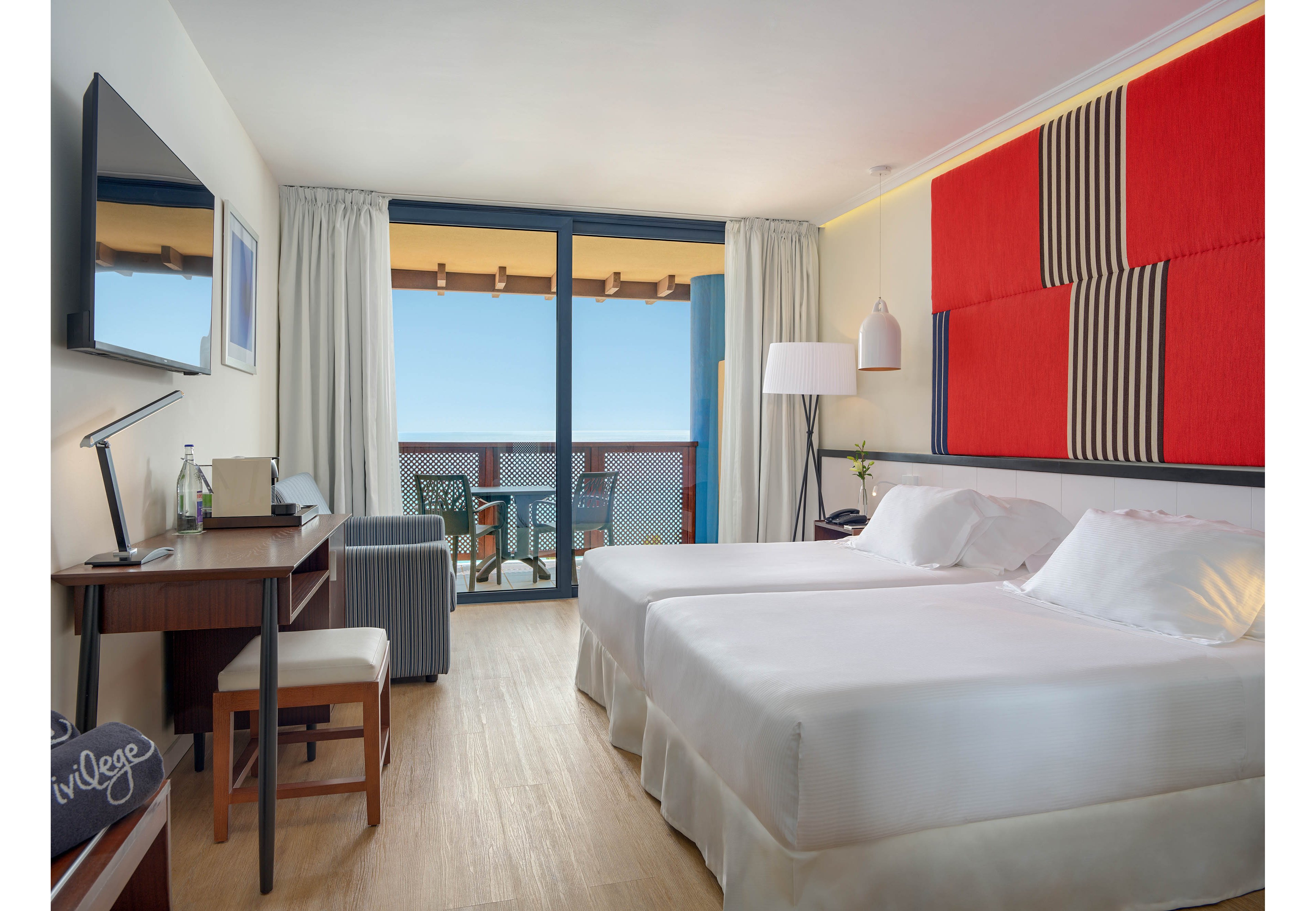 A contemporary hotel room features two neatly made twin beds with white linens and a large red and black abstract wall panel above the headboard. A desk with a chair, flat-screen television, and small seating area with an ottoman sit along one wall. Floor-to-ceiling curtains frame a glass door that opens to a balcony with chairs and a view of the blue ocean and clear sky. Soft neutral tones and clean lines create a calm, minimalist atmosphere.