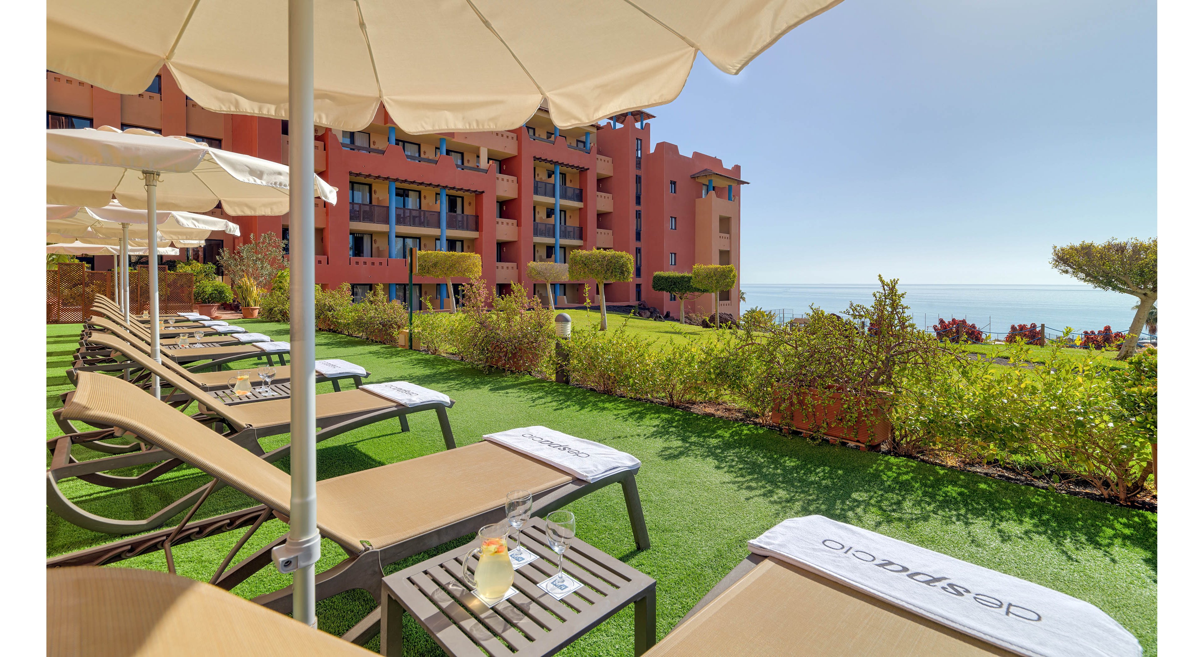 A row of sun loungers with white umbrellas is arranged on a grassy terrace facing the sea. A resort-style building with balconies stands in the background, framed by manicured hedges and topiary trees. A small table between loungers holds a refreshing drink and a glass, suggesting a relaxing vacation atmosphere. The bright daylight and clear sky emphasize a warm, tranquil coastal setting.