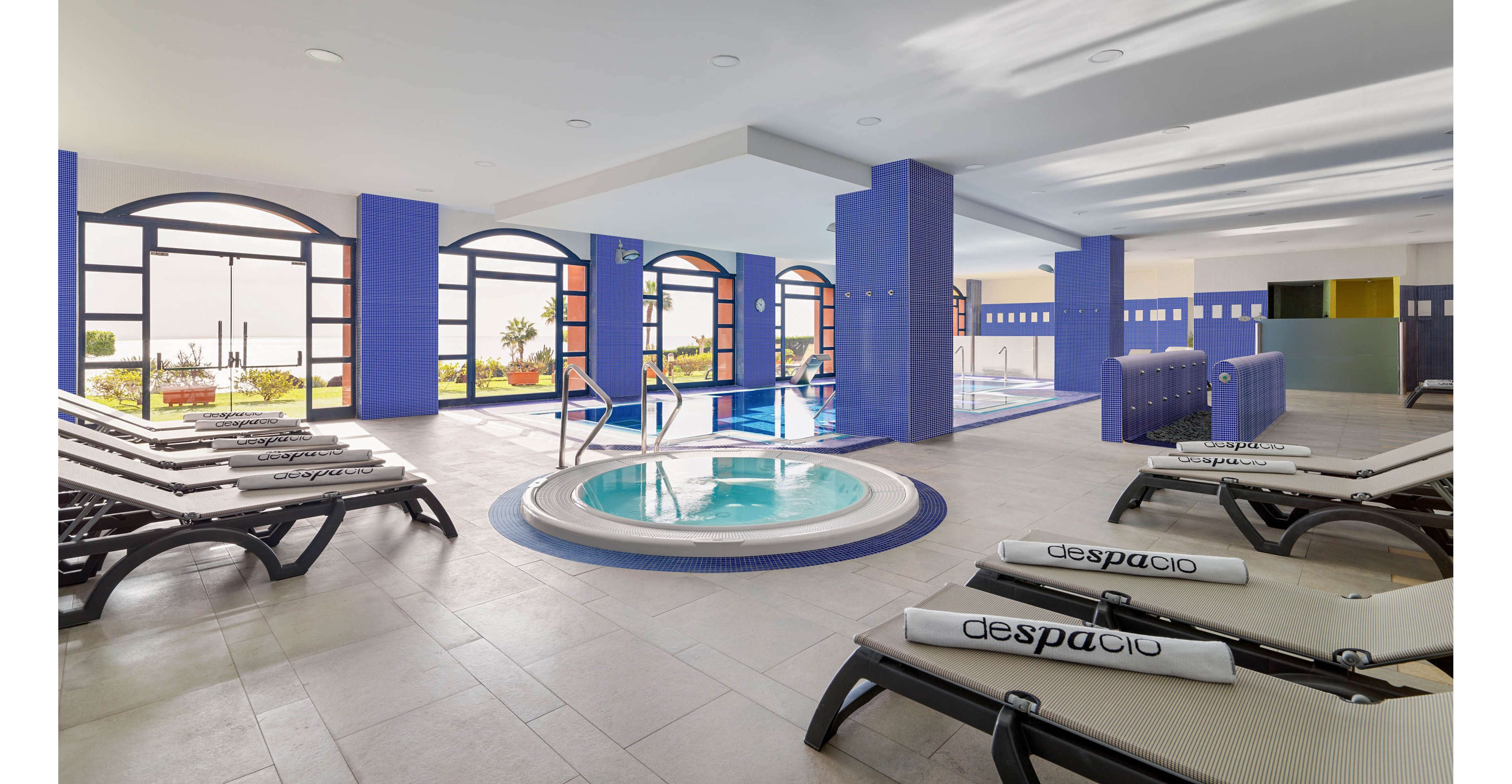 A contemporary indoor spa features a circular jacuzzi at the center surrounded by lounge chairs. Large arched windows line the walls, allowing natural light to brighten the blue and white interior. In the background, a rectangular swimming pool and additional seating areas are visible. Rolled towels are placed on the loungers, creating a clean and inviting wellness atmosphere.