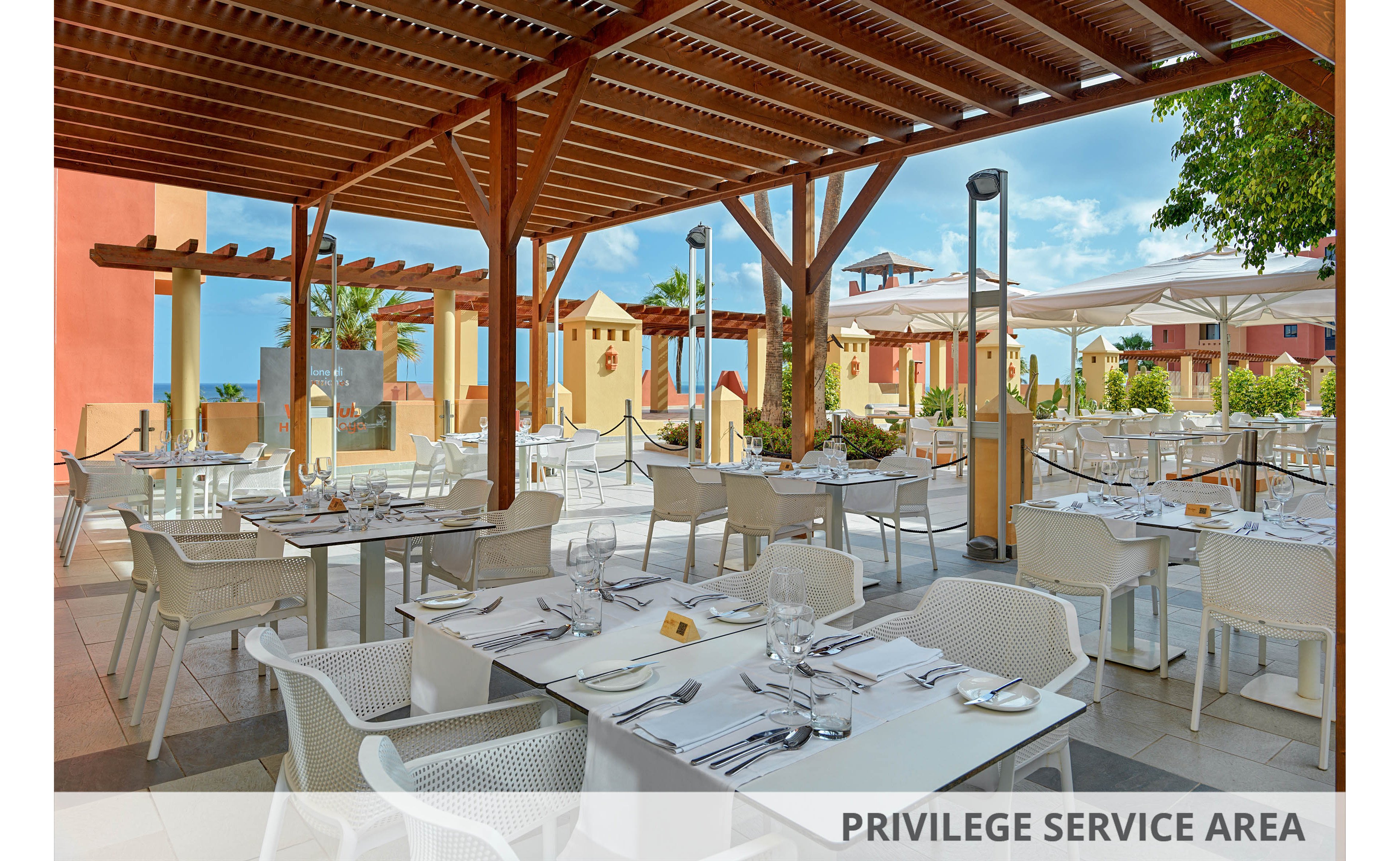 An open-air restaurant terrace is set with neatly arranged white tables and chairs under a wooden pergola. Each table is prepared with glassware, cutlery, and folded napkins, ready for guests. The setting overlooks a bright resort courtyard with colorful buildings, palm trees, and sun umbrellas. The scene suggests a relaxed, upscale dining experience in a warm, coastal climate.