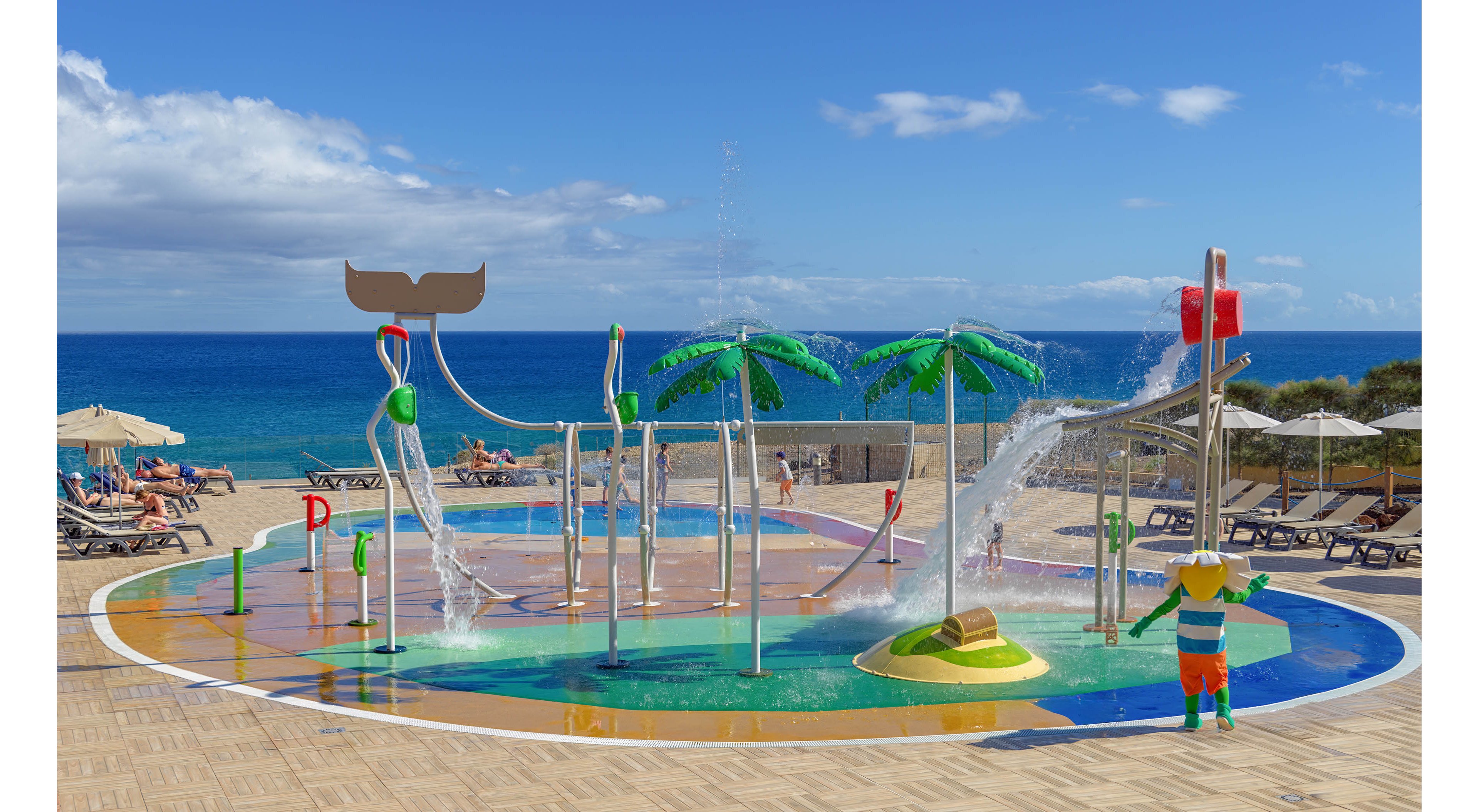 Children play in a colorful splash park near the ocean on a sunny day. The water playground features palm tree fountains, spraying arches, and a large tipping bucket structure. Adults relax on lounge chairs in the background while watching the kids. The bright blue sky and sea create a cheerful, family-friendly holiday atmosphere.