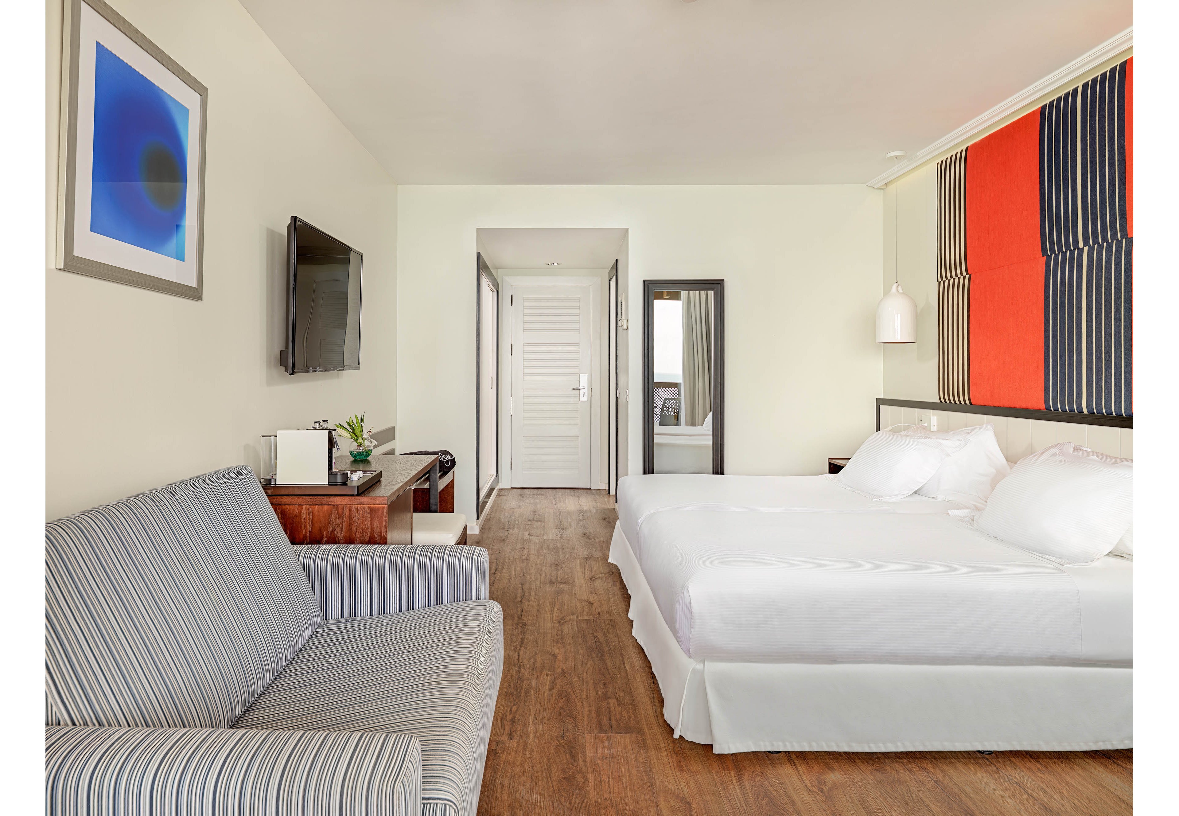 A bright modern hotel room features a large double bed with crisp white linens against a wall with bold striped artwork. A striped sofa sits opposite a flat-screen television mounted on the wall, with a wooden console table and small plant beneath it. The room has light-colored walls, wood-look flooring, and a full-length mirror near the entrance door. Soft lighting and minimal decor create a clean, contemporary atmosphere suitable for travelers.
