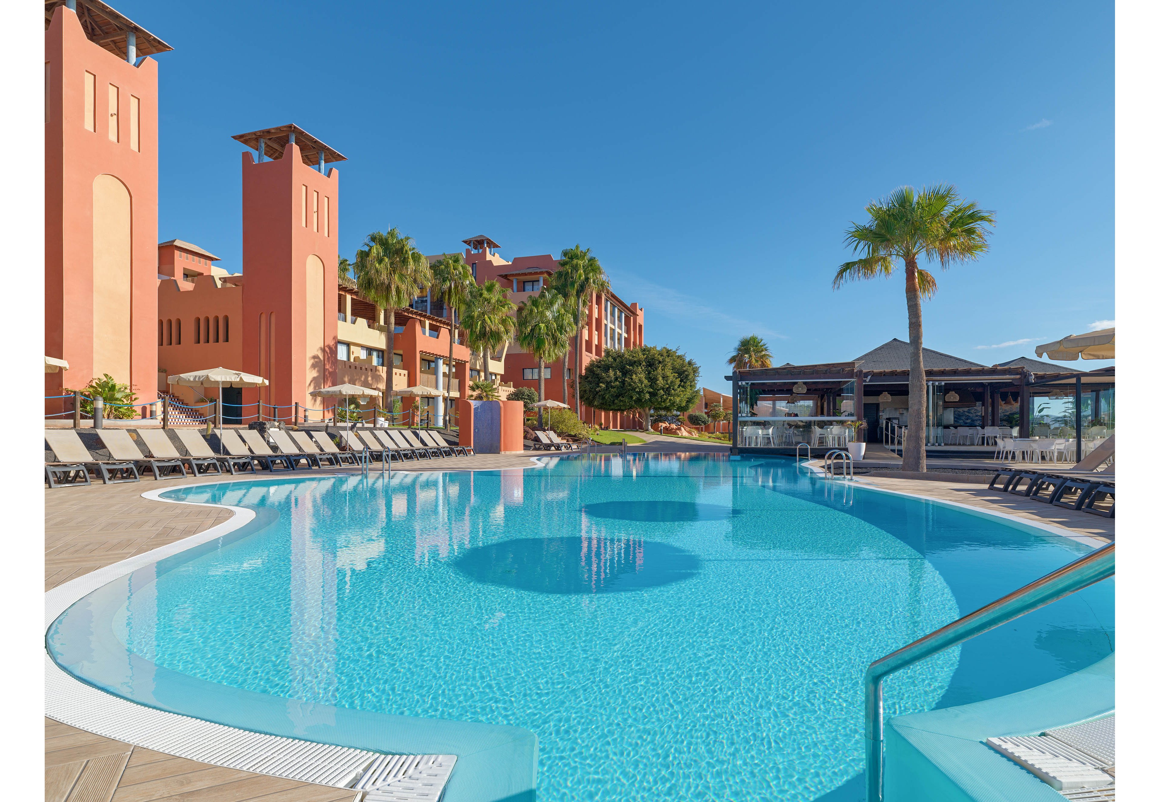 A large, clear blue swimming pool curves gracefully in front of a resort complex. Rows of empty lounge chairs line the poolside under a bright, cloudless sky. Terracotta-colored buildings and tall palm trees create a warm, Mediterranean-style setting. A poolside bar or pavilion stands to the right, adding to the relaxed vacation atmosphere.