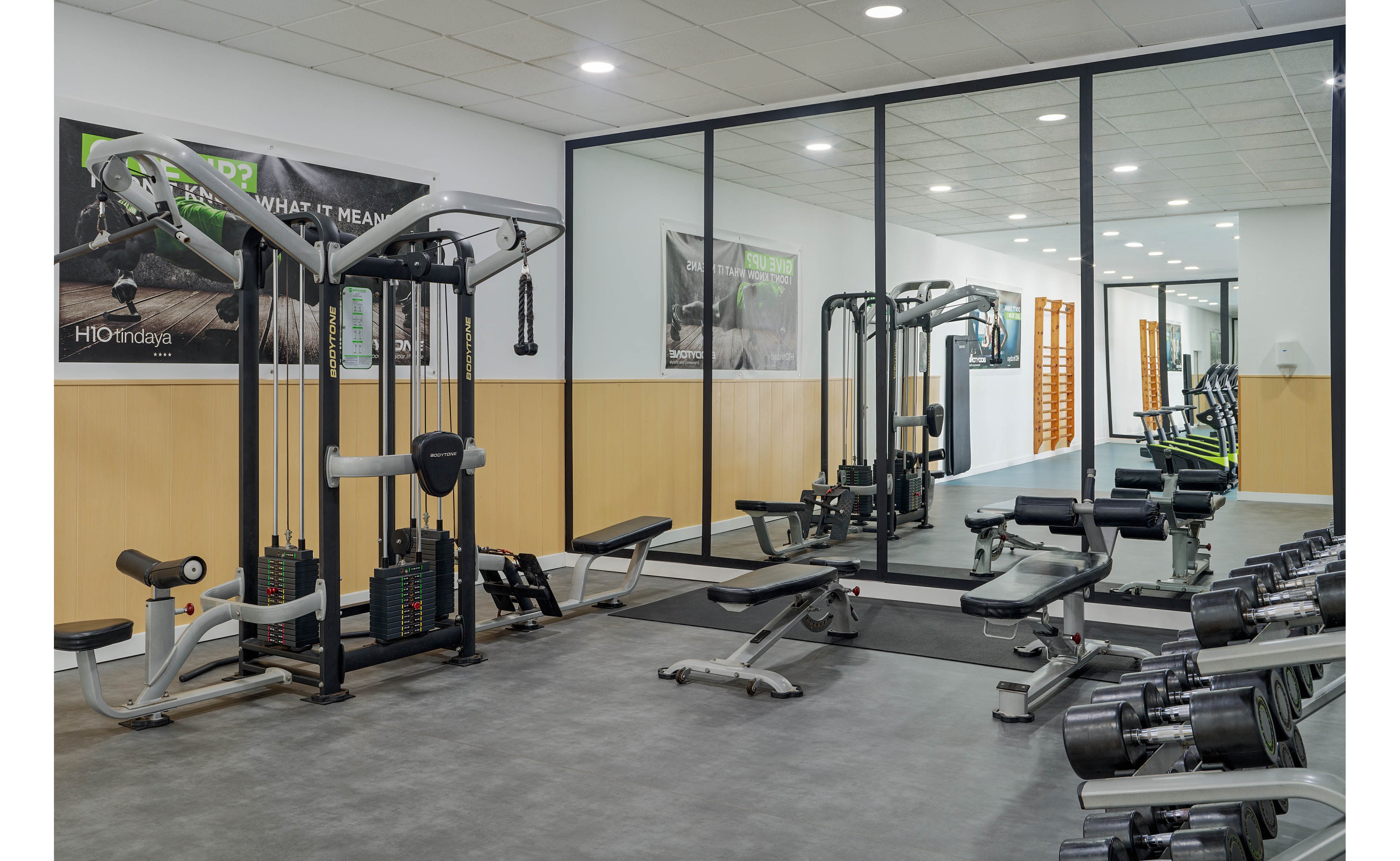 A modern fitness room features various strength training machines and free weights neatly arranged. The space includes cable machines, benches, and dumbbells facing a large mirrored wall. Bright overhead lighting and light-colored wall panels create a clean, professional atmosphere. Motivational posters are visible on the walls, enhancing the contemporary gym setting.