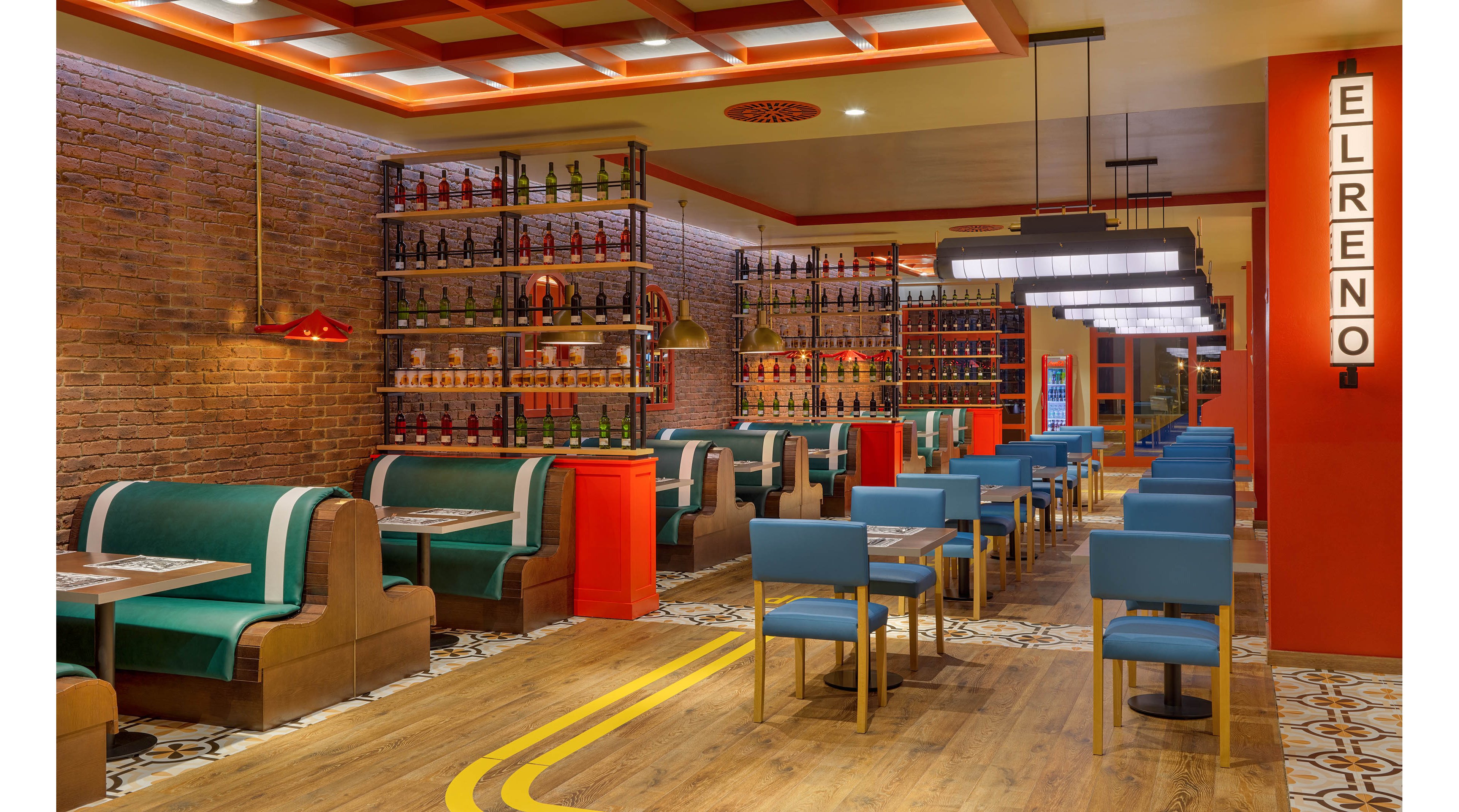 A vibrant restaurant interior features teal and brown upholstered booths arranged along a warm wooden floor. Rows of blue chairs and tables line the opposite side, creating a symmetrical dining layout. Shelves filled with bottles decorate the exposed brick wall, adding visual interest and color. Bright orange accents, patterned floor tiles, and contemporary pendant lights contribute to a modern, inviting atmosphere, with a vertical sign reading "EL RENO" visible on the right.