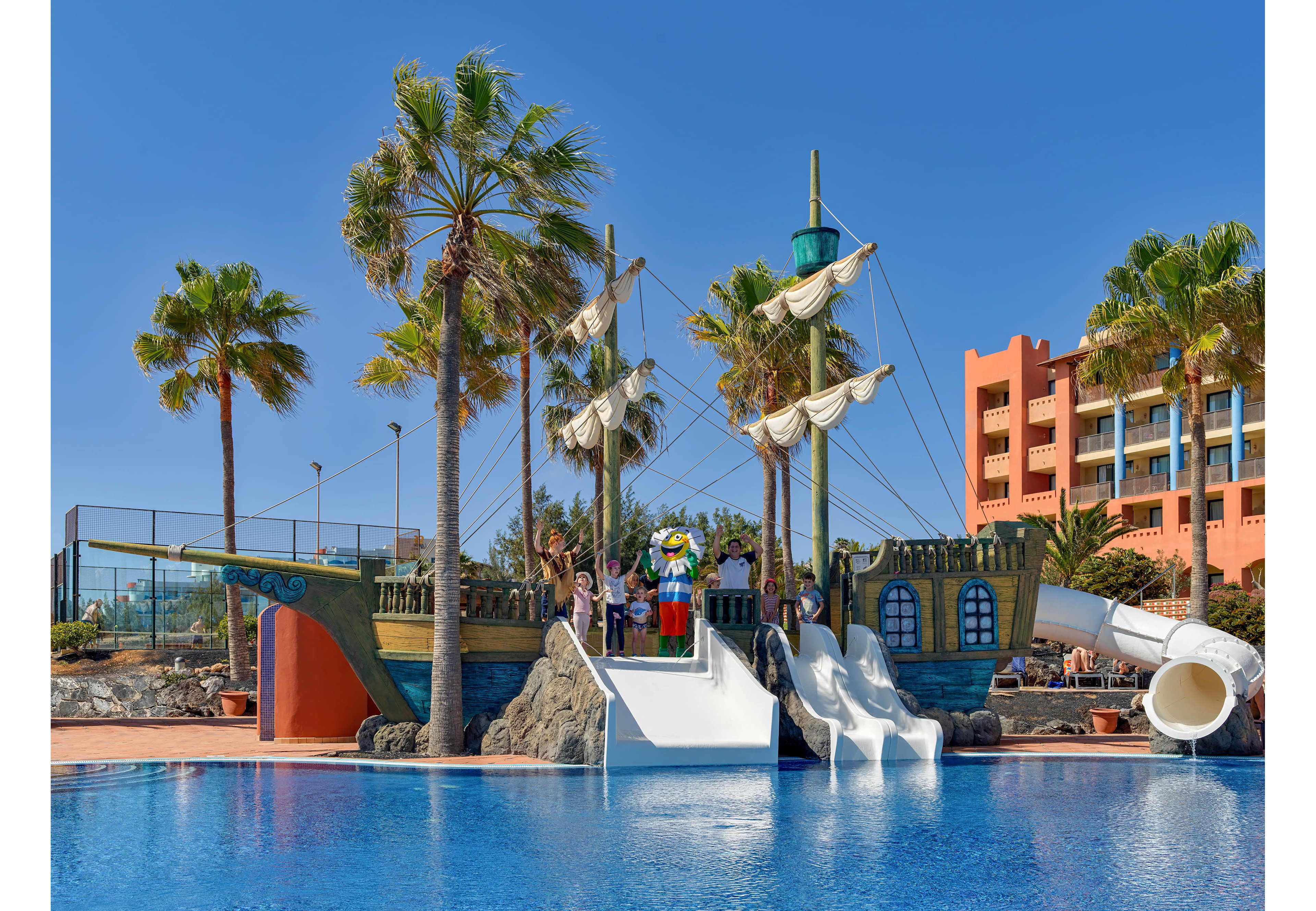 A colorful pirate ship themed play structure stands beside a bright blue swimming pool. Children are gathered on the deck and using multiple white water slides that descend into the pool. Tall palm trees sway around the attraction, and a resort building with balconies is visible in the background. The clear sky and vivid colors suggest a sunny, warm vacation setting.