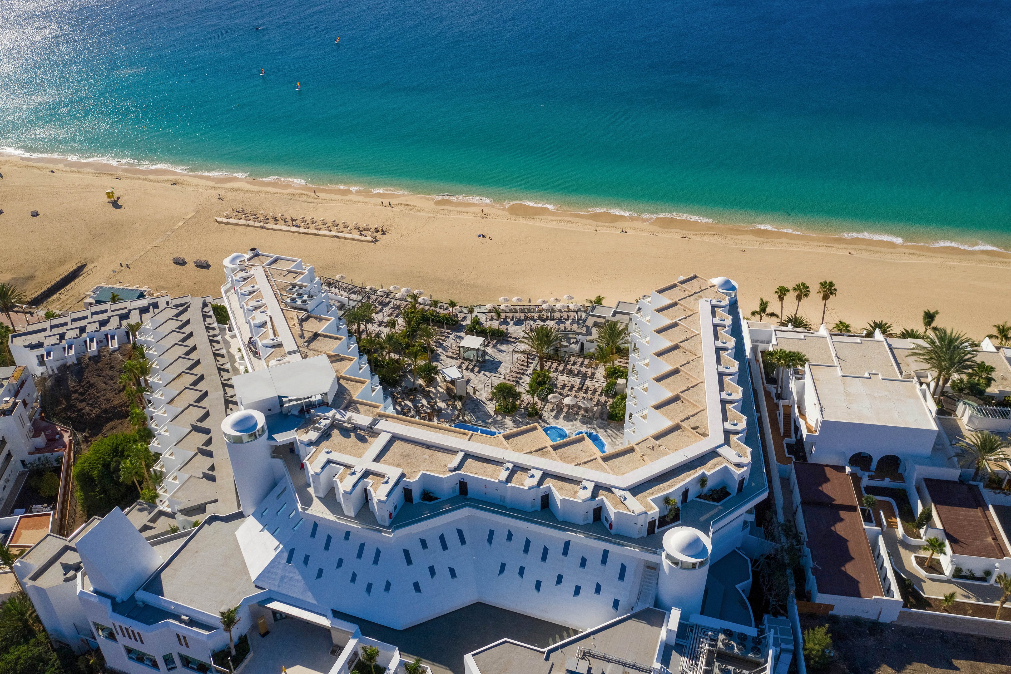 The image shows a large white resort complex situated directly on a sandy beach, with clear turquoise water in the background. Rows of lounge chairs and umbrellas are visible along the shoreline, and palm trees are scattered throughout the property. The architecture features modern, angular lines and multiple levels, creating a striking visual contrast with the natural surroundings. No visible text or numbers are present in the image.