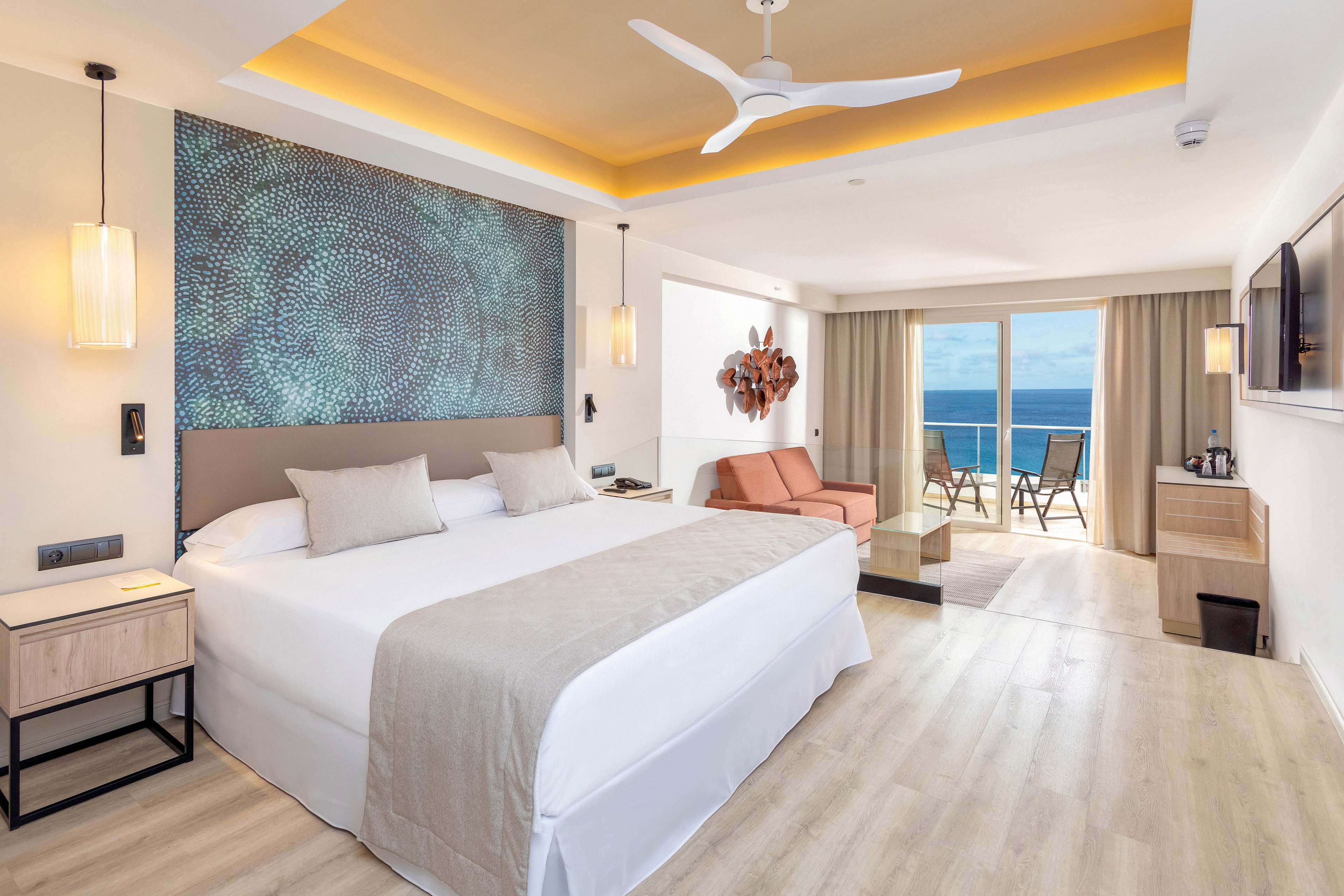 A spacious hotel room features a king-sized bed, neutral decor, and contemporary furnishings. The setting includes a cozy seating area, wall art, and a balcony overlooking the ocean. Warm lighting and a ceiling fan enhance the relaxing atmosphere, while natural light fills the space. The room is styled in soft beige and white tones, creating a tranquil mood.