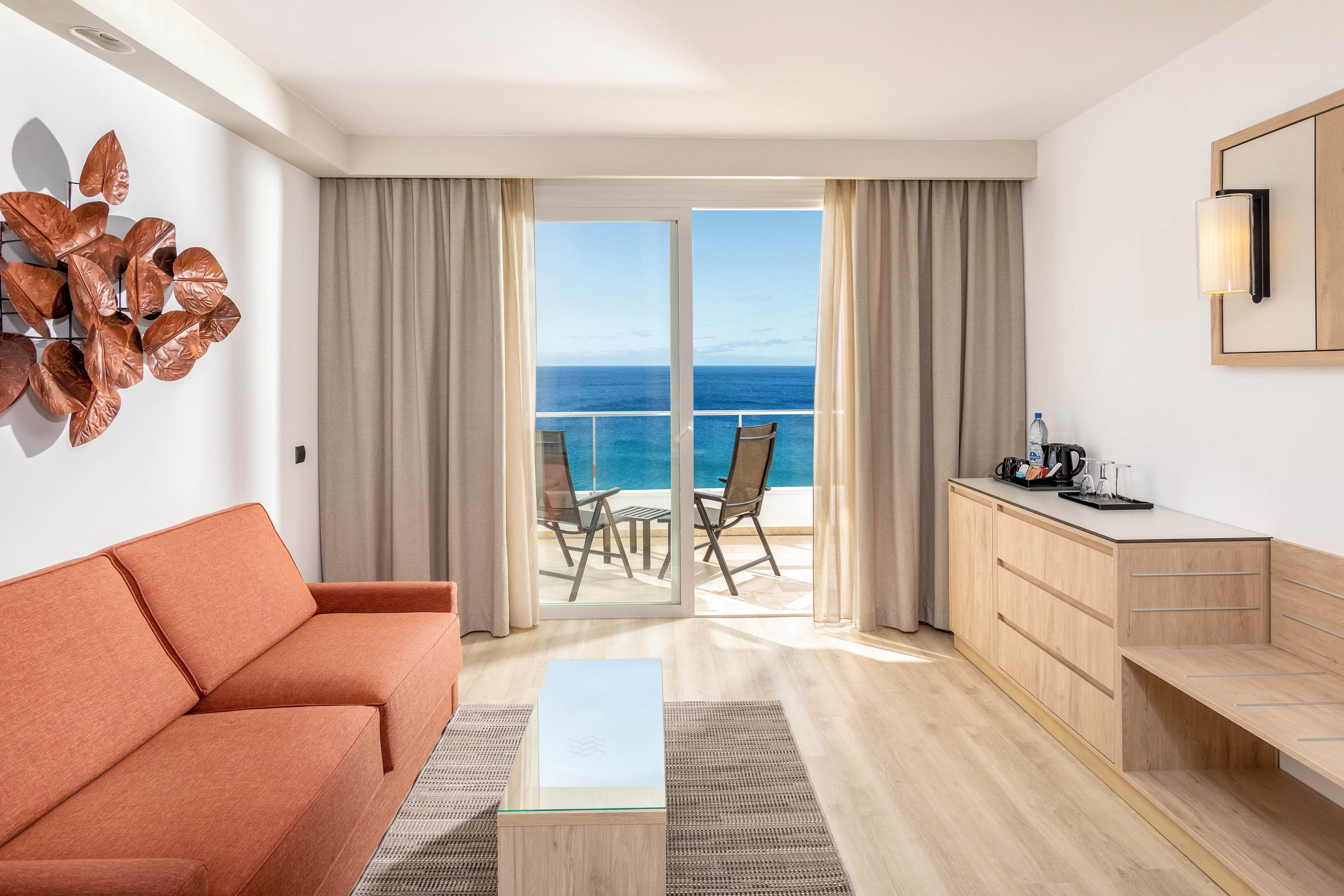 A contemporary hotel room features a burnt orange sofa, glass coffee table, and light wood furnishings. Large sliding doors open to a balcony with two chairs overlooking a vibrant blue ocean. The space is bright and airy, with neutral tones and minimalist decor, including a wall sculpture and beverage station.