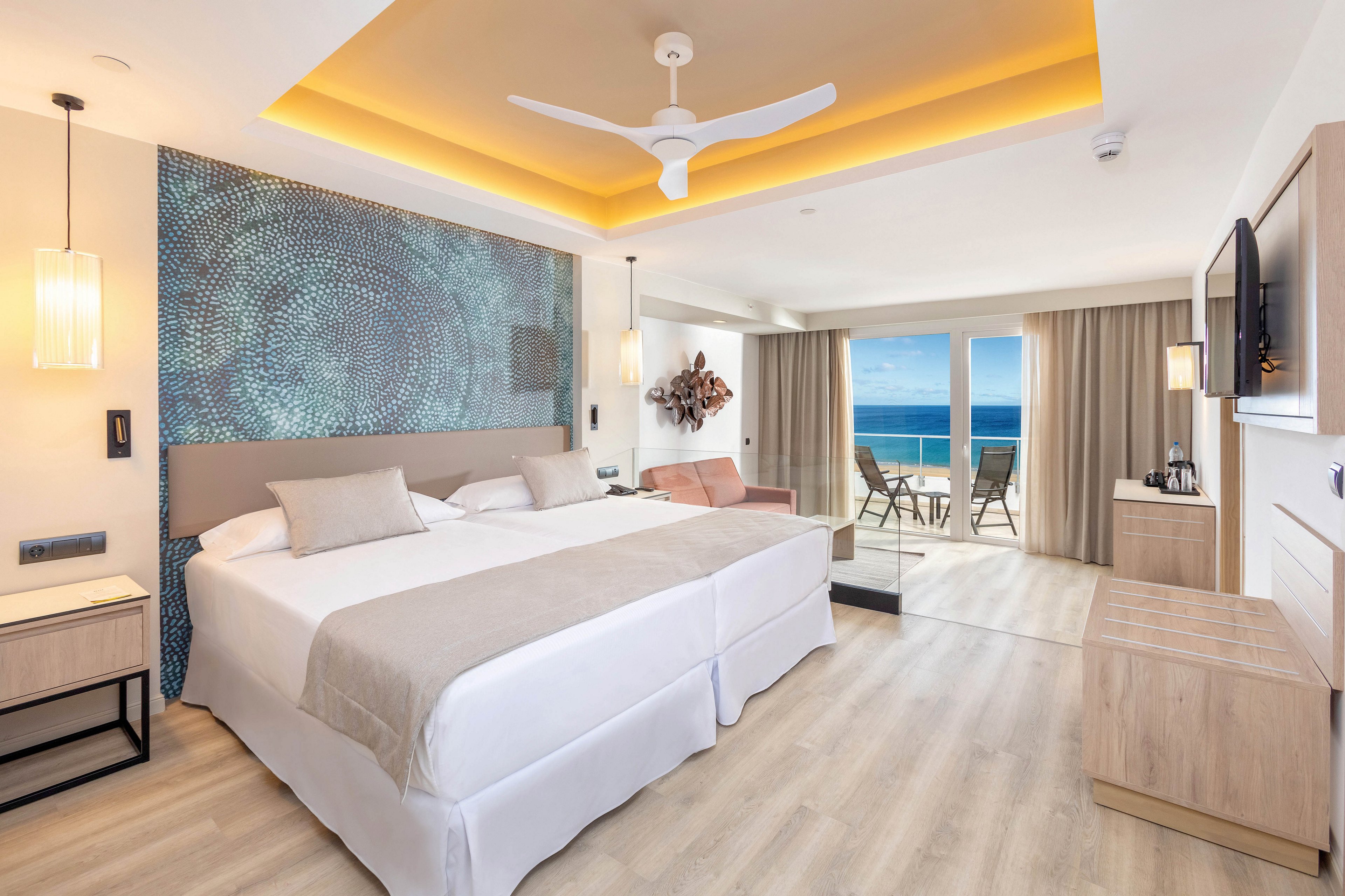 A spacious, contemporary hotel room features two large beds with neutral bedding, a textured accent wall, and modern lighting. The room opens to a private balcony overlooking the ocean, with outdoor seating visible through glass doors. Light wood flooring, minimalist furniture, and soft beige tones create a calm, inviting atmosphere.