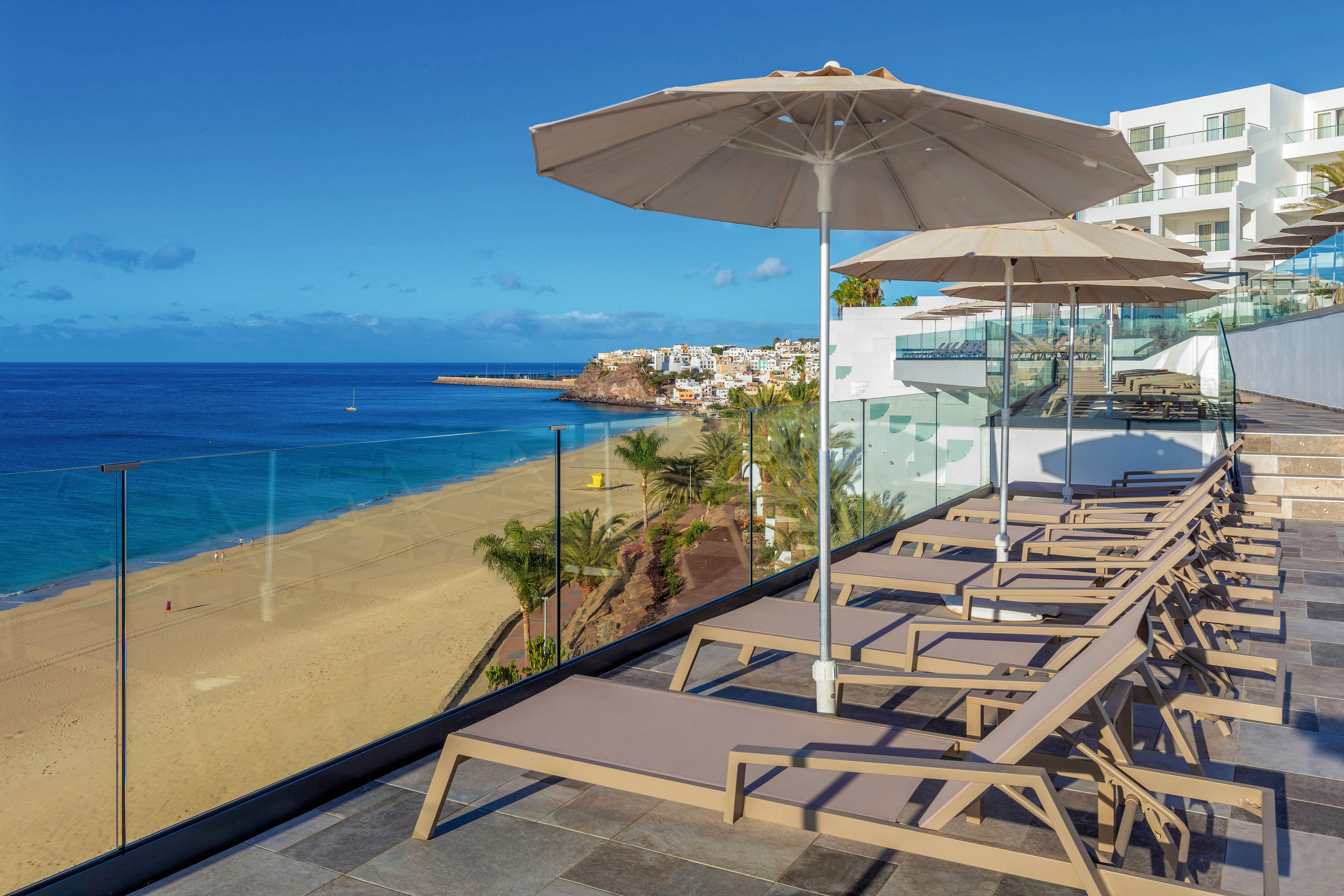 A contemporary terrace overlooks a sandy beach and clear blue ocean, featuring several lounge chairs and large umbrellas. The setting is sunny and tranquil, with glass railings providing unobstructed views of the coastline and nearby white buildings. The mood is relaxed and inviting, ideal for leisure and vacation.