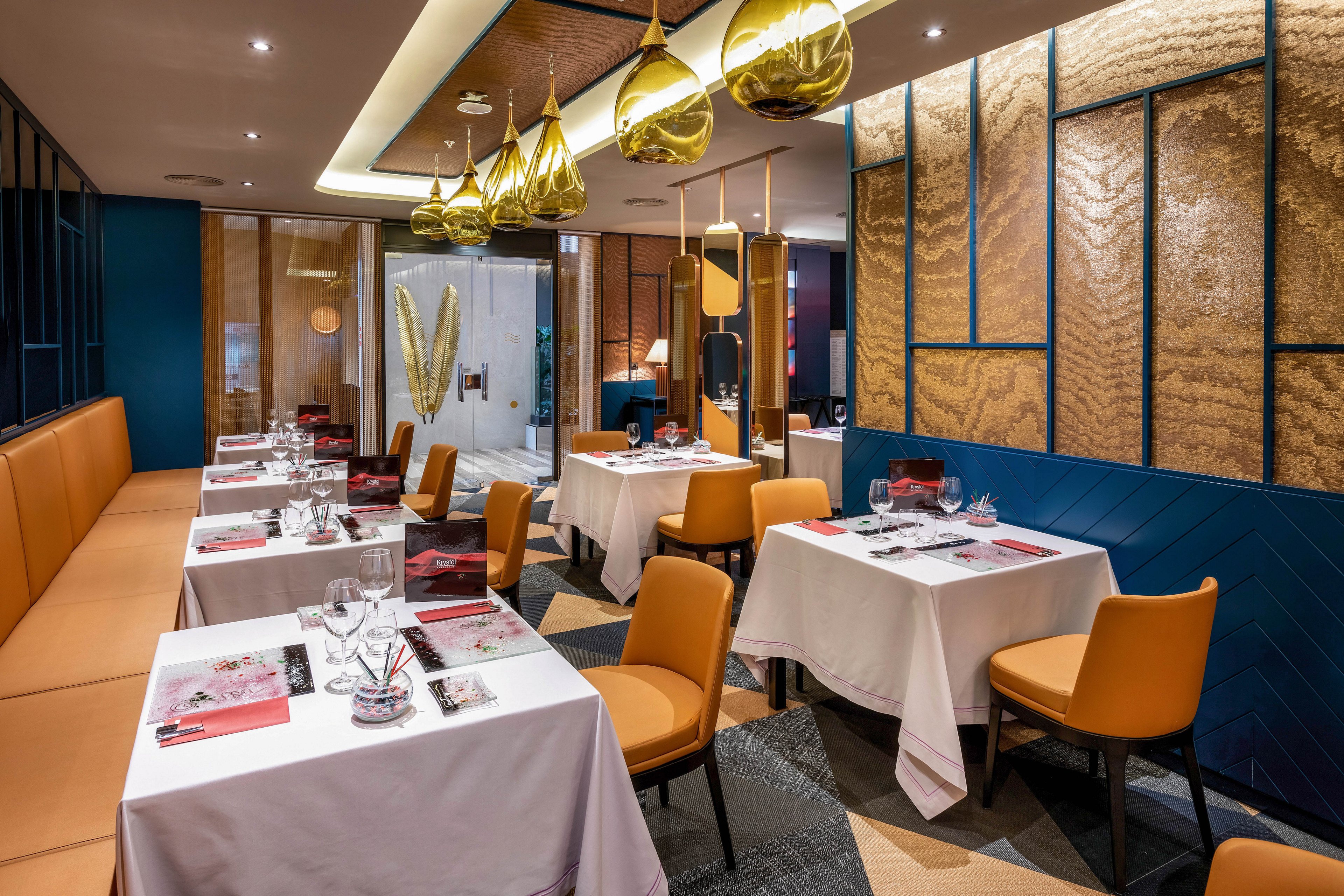 The image features a contemporary restaurant dining area with neatly arranged tables covered in white linens. Warm brown leather chairs and banquette seating line the space, complemented by gold pendant lights and textured wall panels. The setting is well-lit, stylish, and inviting, with a sophisticated ambiance and a neutral palette. No visible text or numeric values are present in the scene.