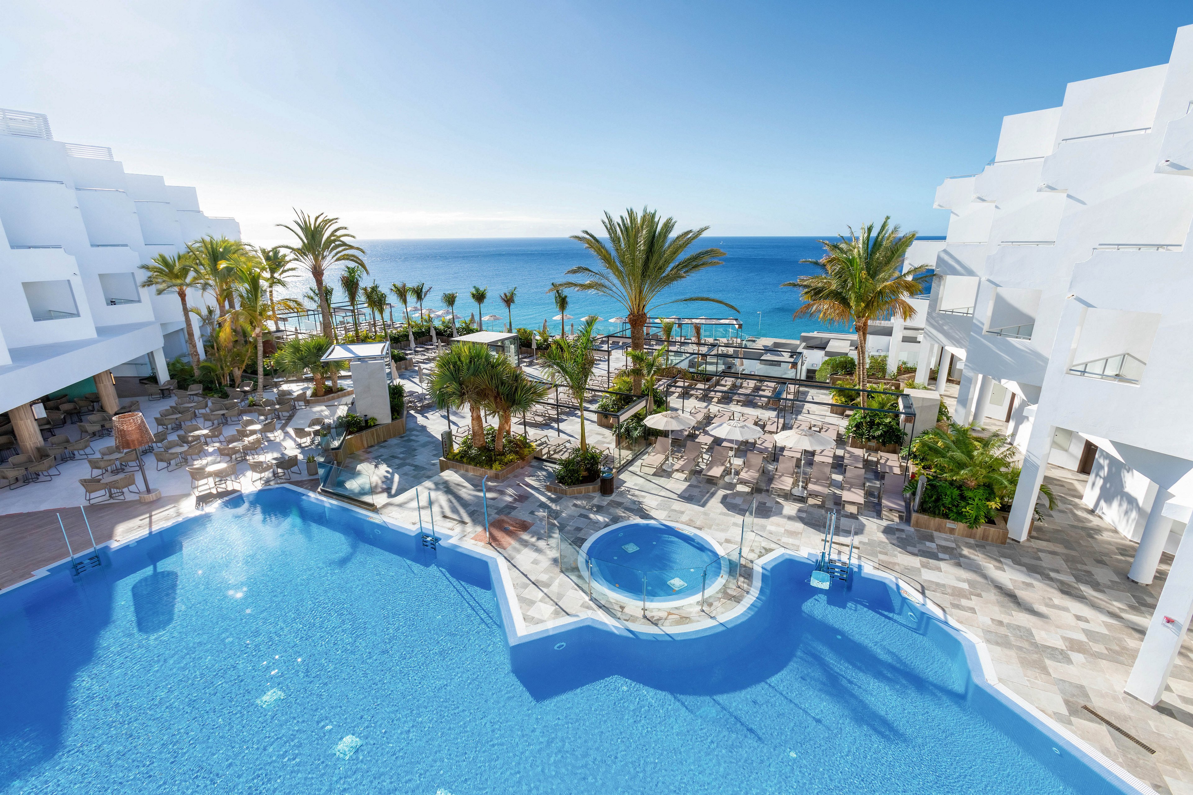 A modern resort features a large swimming pool and a circular hot tub surrounded by palm trees and sun loungers. The setting overlooks a clear blue ocean under a bright sky, with white geometric buildings framing the scene. The atmosphere is tranquil and inviting, ideal for relaxation and leisure by the sea.