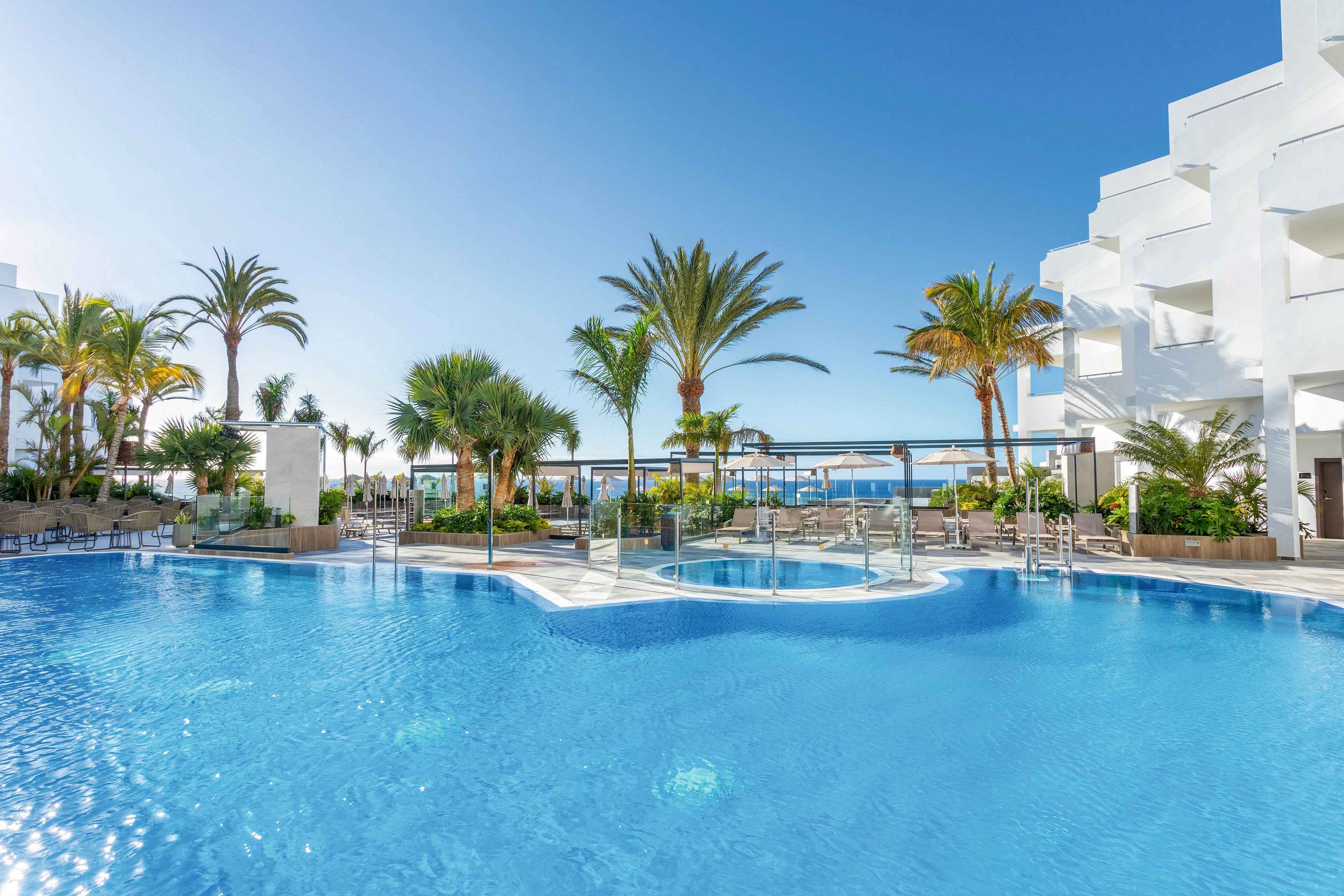 A spacious outdoor swimming pool is featured in a contemporary resort setting, surrounded by white buildings and lush palm trees. The scene is bright and sunny, with clear blue water reflecting the sky. Lounge chairs and shaded areas are visible, creating a relaxing atmosphere for guests. No visible text or numeric values are present in the image.