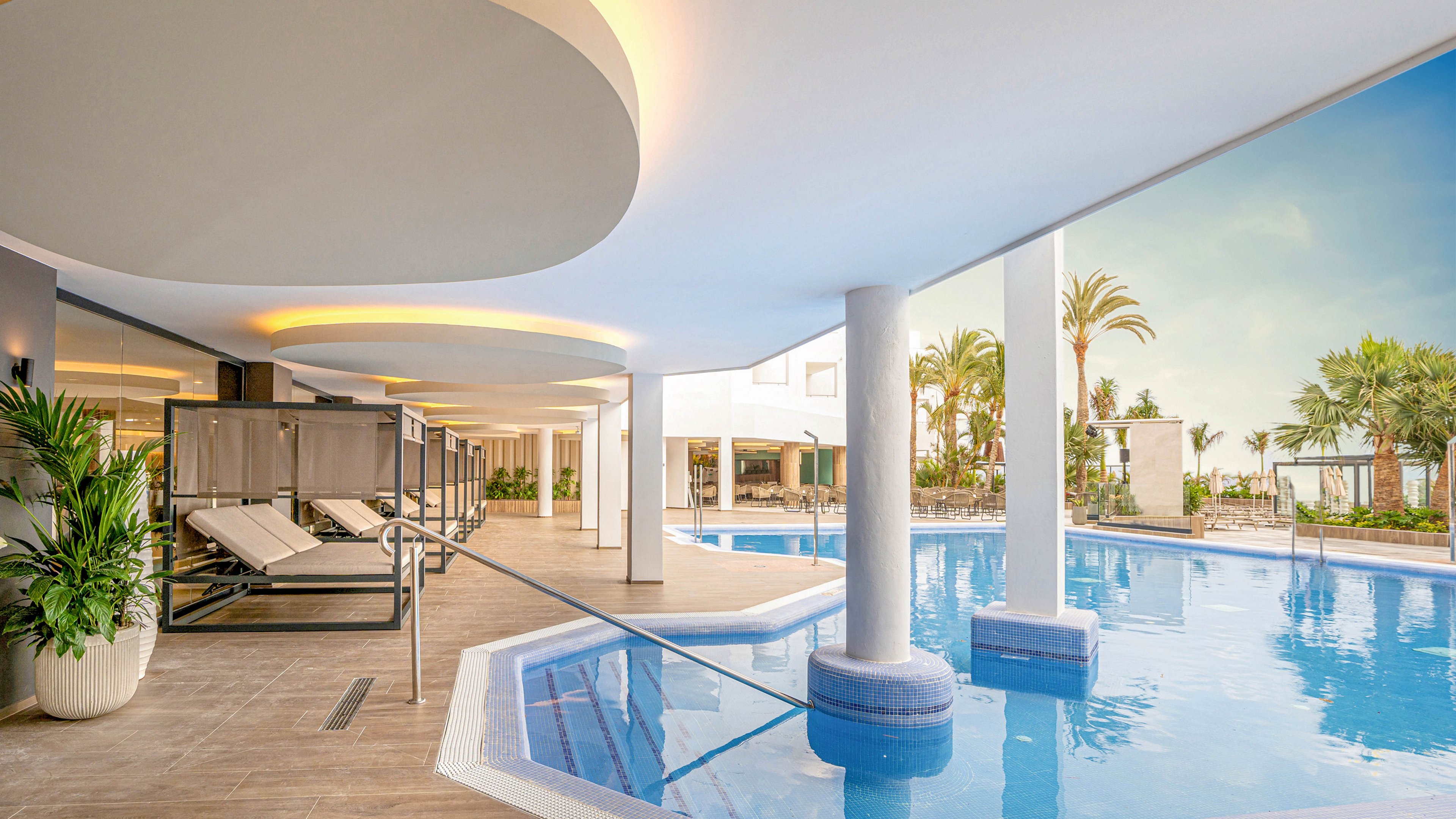 The image features a contemporary pool space blending indoor and outdoor elements, with sleek lounge beds positioned beside the water. Large columns and curved ceiling details create a stylish, open atmosphere. Palm trees and lush greenery are visible outside, enhancing the relaxing mood. The scene is brightly lit with natural and artificial light, emphasizing a tranquil, resort-like ambiance.