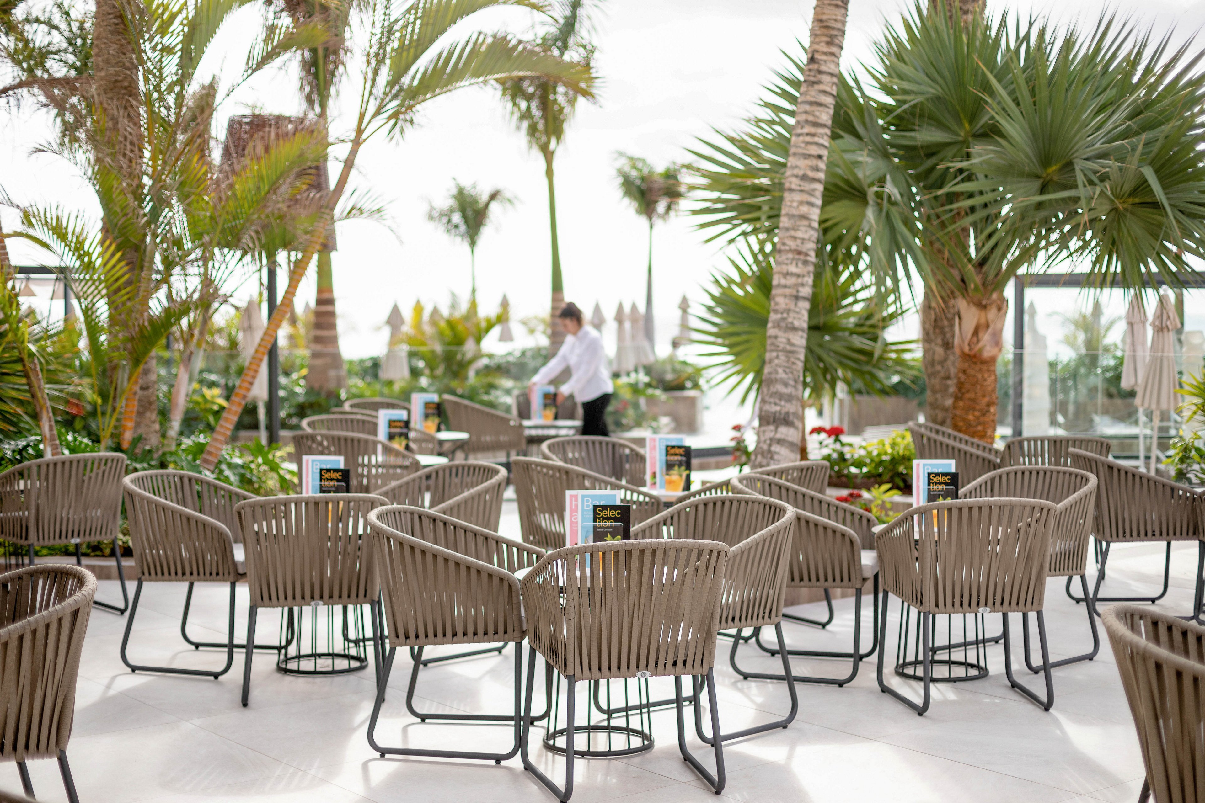 A sunlit outdoor cafe features modern woven chairs and tables arranged among lush palm trees. A person in white attire is seen in the background, possibly a staff member preparing the area. Menus or signs are visible on each table, adding to the organized and inviting atmosphere. The setting suggests a relaxed, tropical vibe with natural greenery and bright daylight.