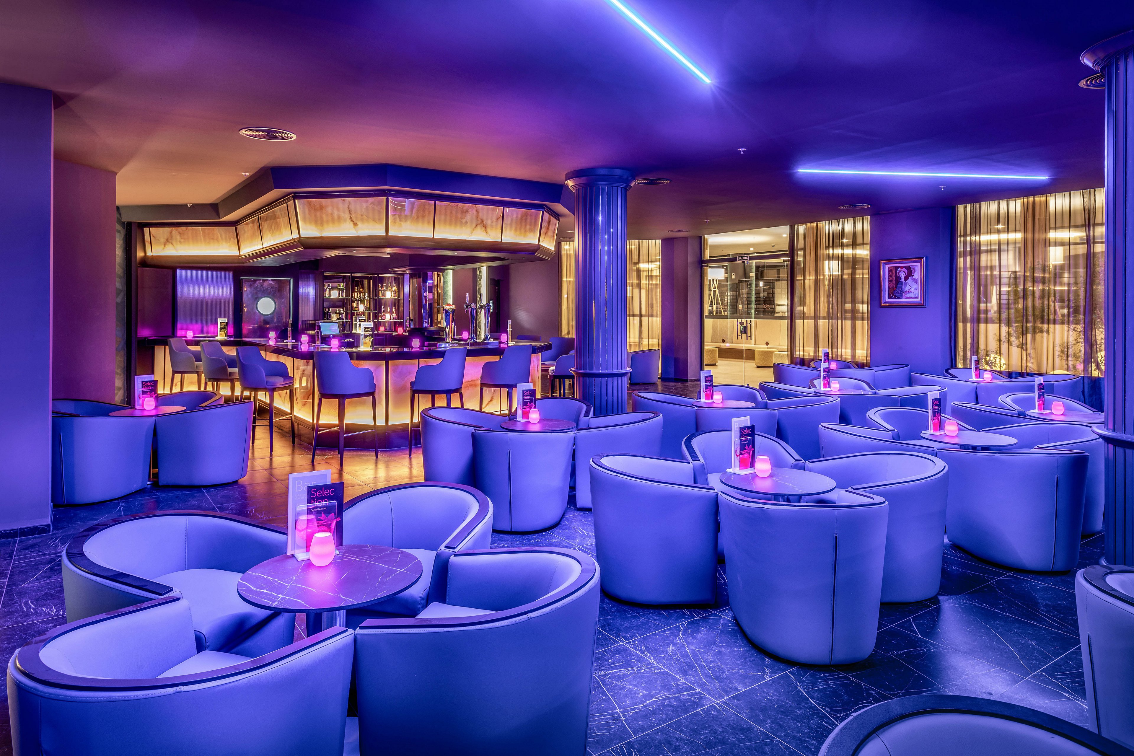 A contemporary lounge bar features curved armchairs and round tables, illuminated by vibrant neon purple and pink lighting. The bar area is visible in the background, with high stools and a sleek counter. The setting is stylish and inviting, with a futuristic ambiance and glowing table lights. No visible text or numeric values are present in the image.