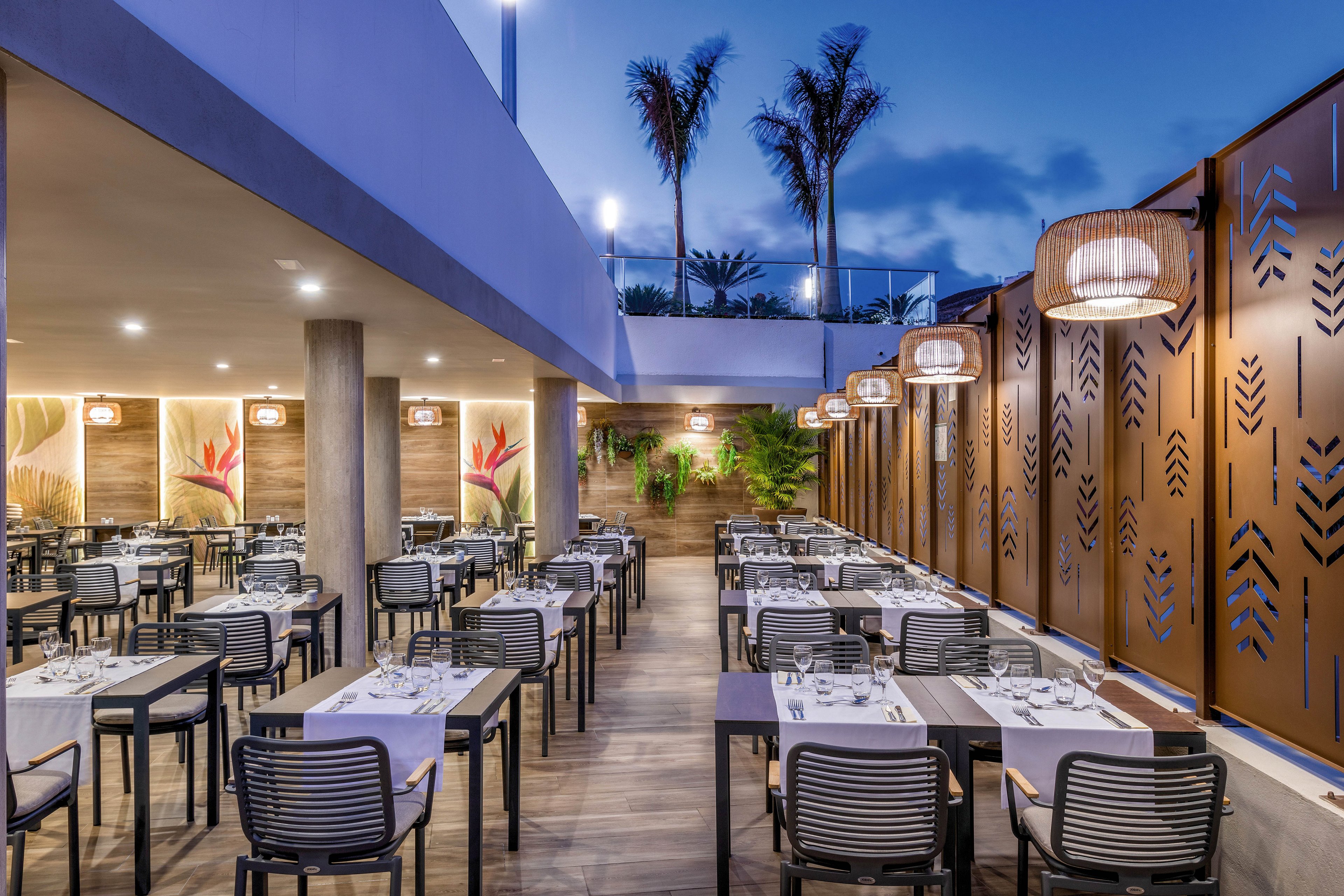 The image features a contemporary outdoor restaurant with neatly arranged tables and chairs, set for dinner service. The setting is illuminated by stylish hanging lights and surrounded by decorative panels and tropical plants, creating a welcoming ambiance. The sky is transitioning to evening, adding a serene blue tone to the scene. No visible text or numeric values are present in the image.