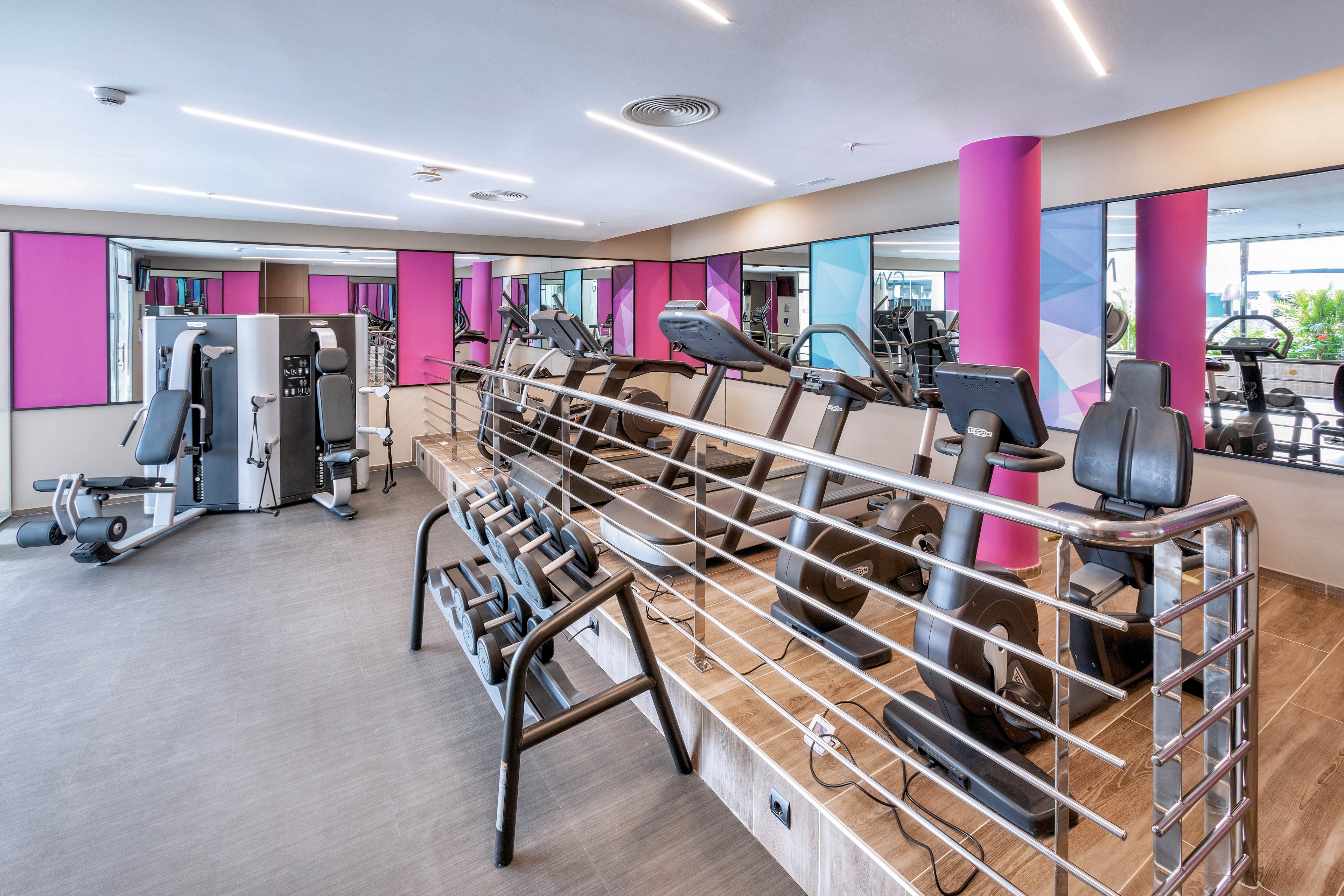 A contemporary gym interior featuring various exercise machines, including treadmills, stationary bikes, and strength training equipment. The space is brightly lit with purple accent columns and mirrors lining the walls, creating a spacious and energetic atmosphere. No people are present, and the gym appears clean and organized, with a neutral palette and pops of color.