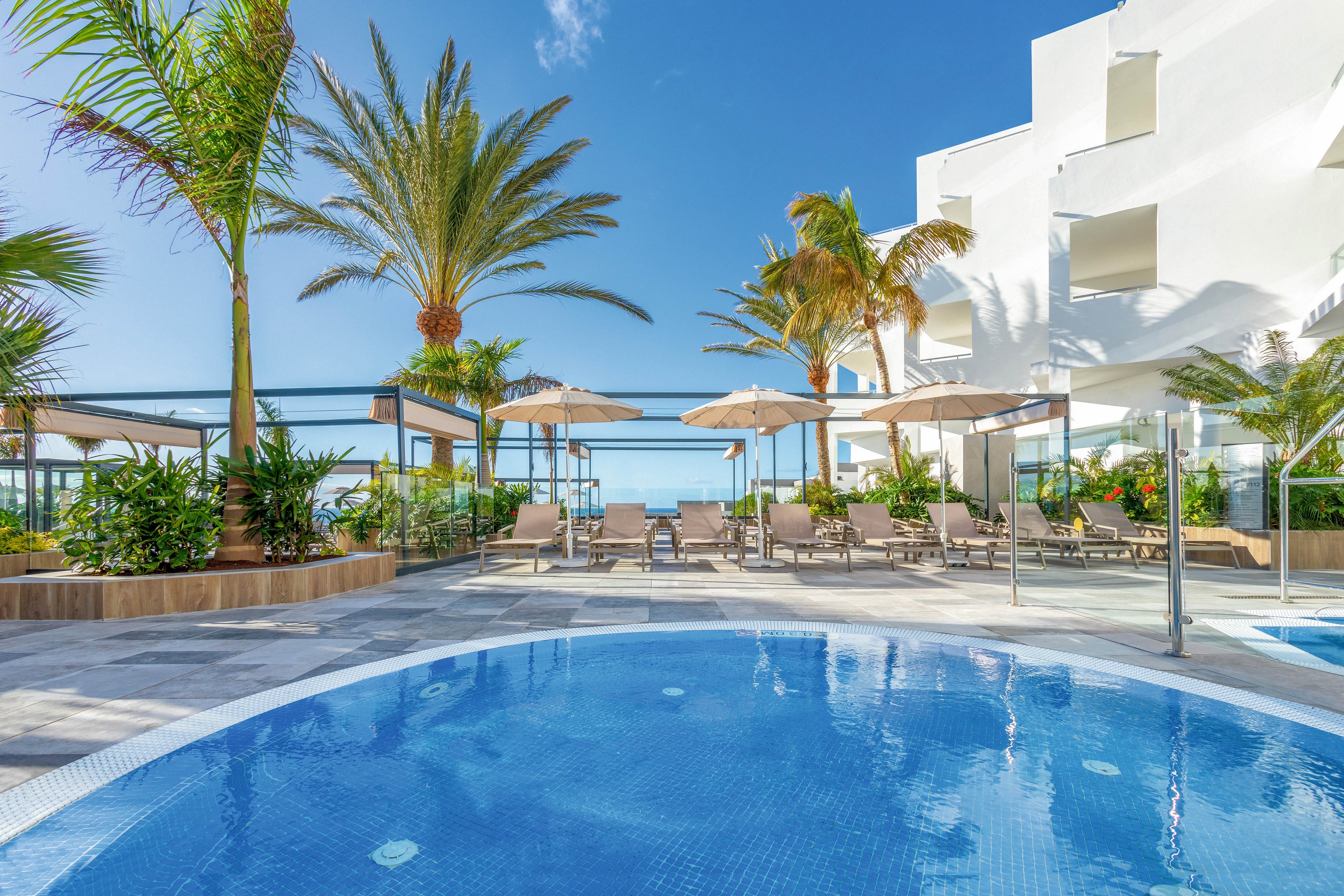 A contemporary outdoor swimming pool is surrounded by palm trees and sun loungers under beige umbrellas. The setting features a clear blue sky and a white modern building, evoking a relaxing vacation atmosphere. The pool’s water is vibrant blue, and the area is landscaped with tropical plants. No visible text or numbers are present in the image.