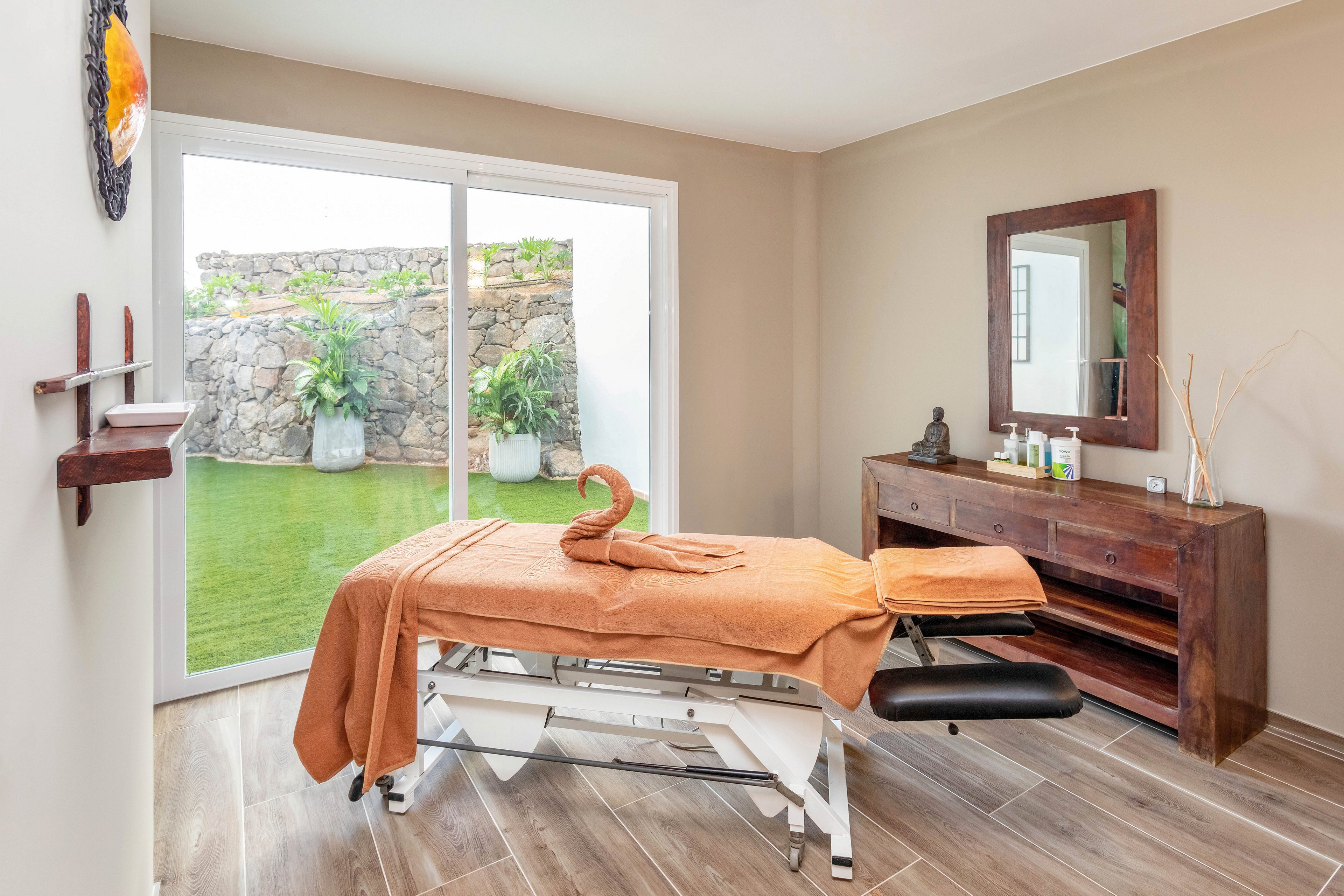 A tranquil spa treatment room features a massage table covered with an orange towel, artfully folded into a swan shape. The space is bright with natural light from large sliding glass doors that open to a small garden with stone walls and potted plants. The room includes wooden furniture, a mirror, and neatly arranged spa essentials, creating a calm and inviting atmosphere.