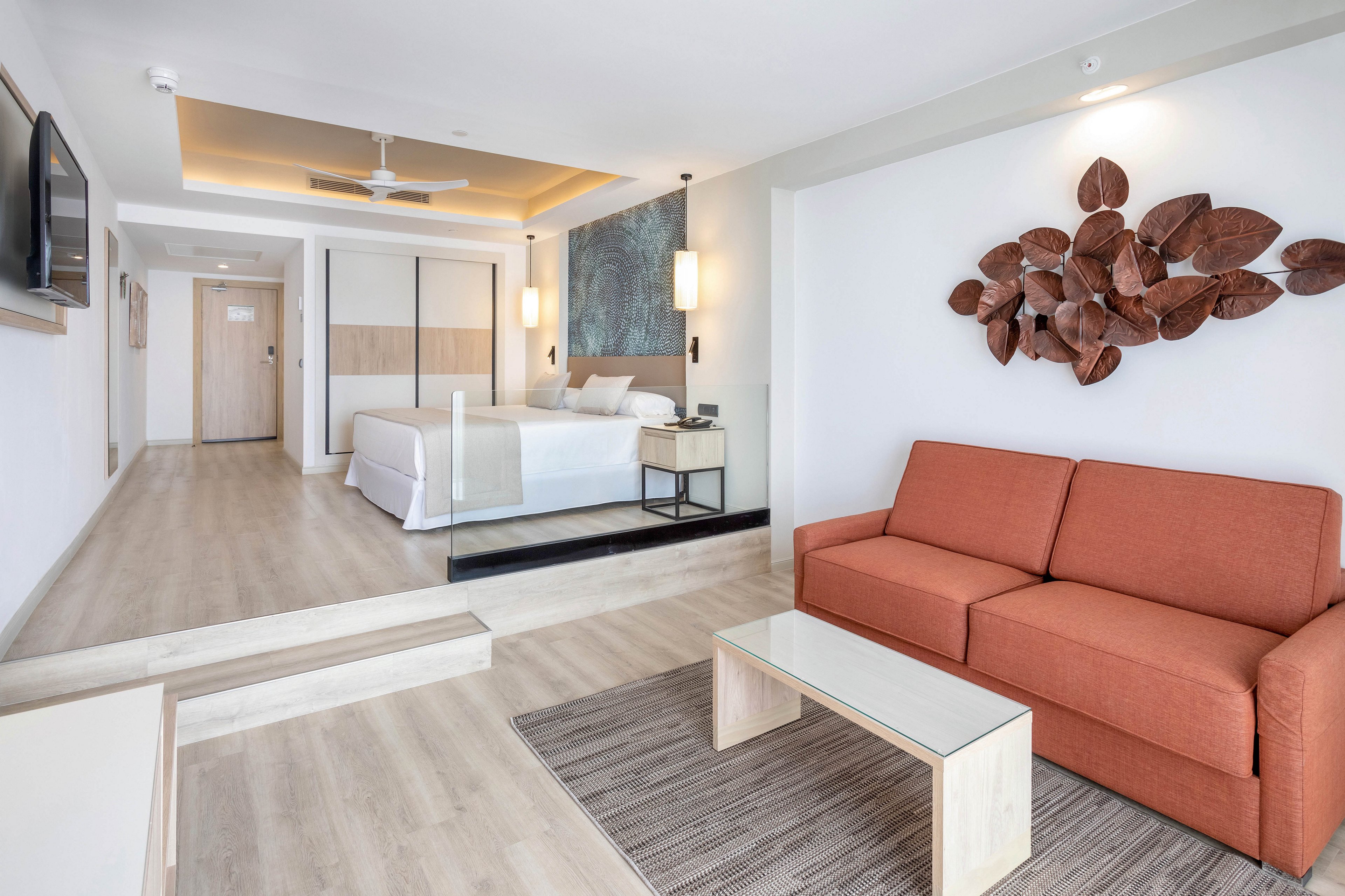 A spacious hotel suite features a king-sized bed, orange sofa, and glass coffee table. The room is decorated with neutral tones and contemporary art, including a leaf-themed wall sculpture. Soft lighting and clean lines create a calm, inviting atmosphere. The suite includes a flat-screen TV and a ceiling fan, with light wood flooring throughout.